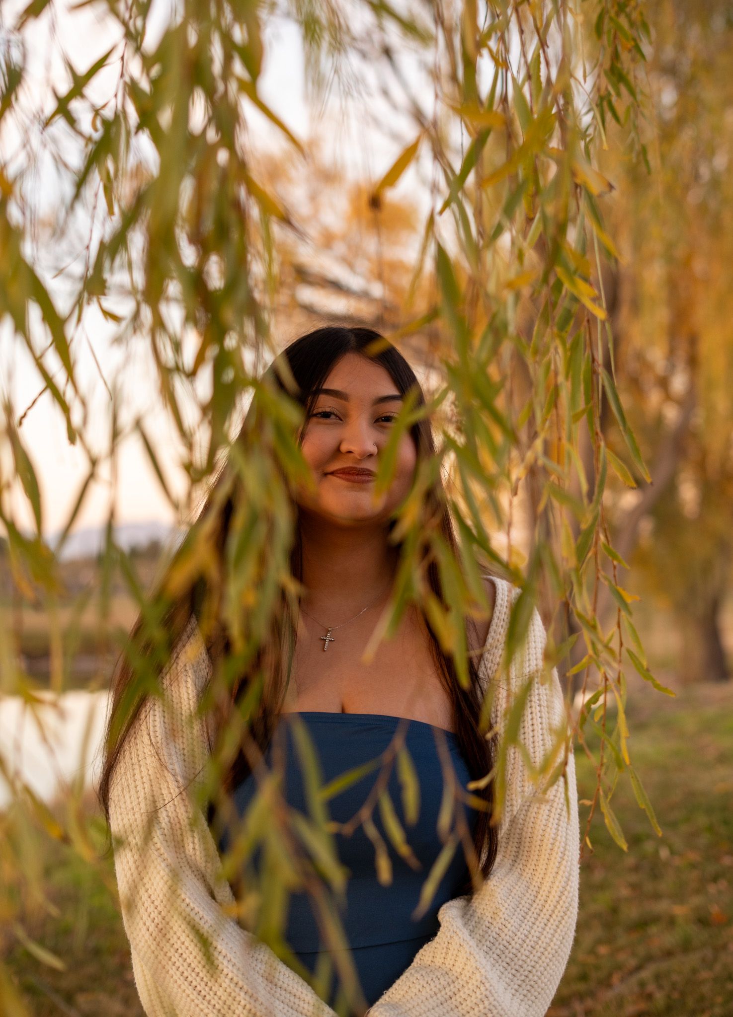Portrait through willow branches of a woman in blue dress and white sweater at golden hour.