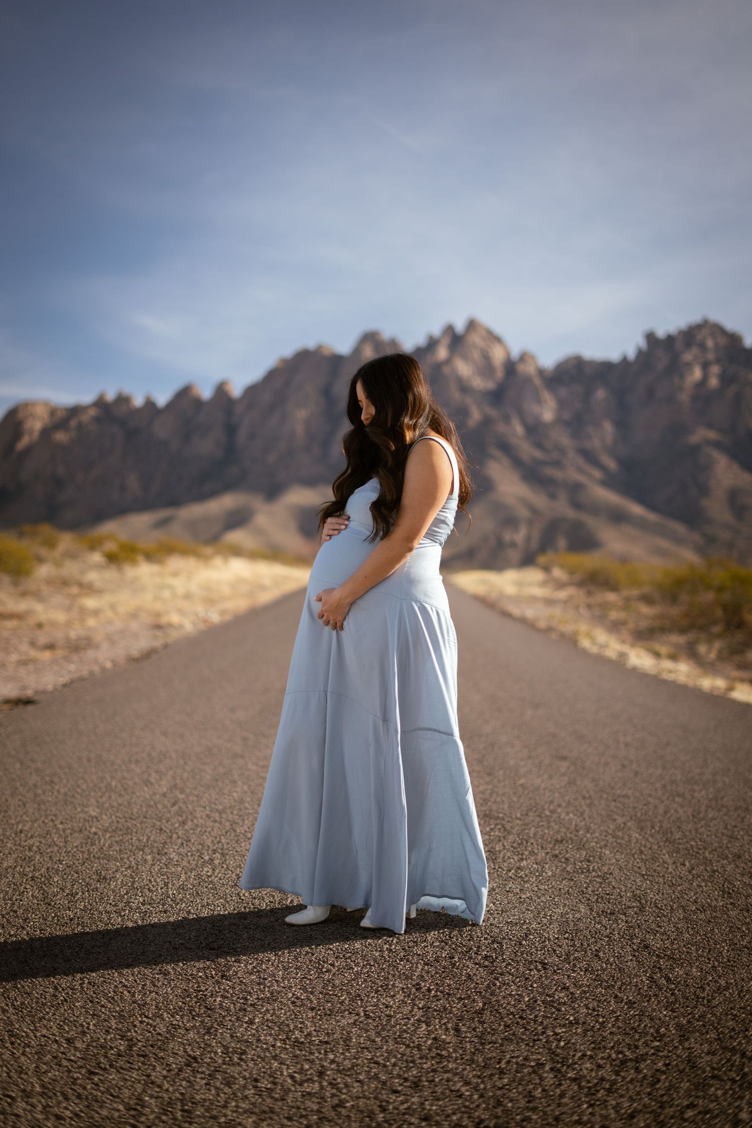 A pregnant woman in a flowing light blue dress stands on an empty desert road with rugged mountains in the background.