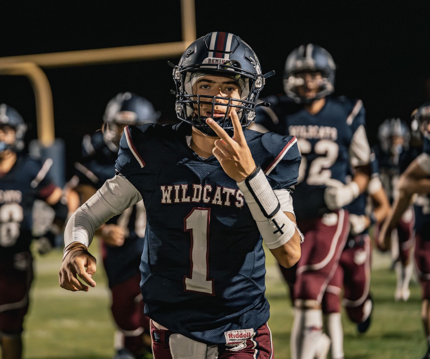 High school football player in navy uniform runs with ball during nighttime game on illuminated field.