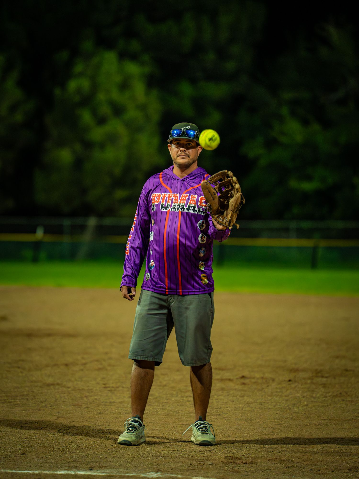 Softball player in purple jersey and grey shorts prepares to catch a yellow ball on a dirt field.