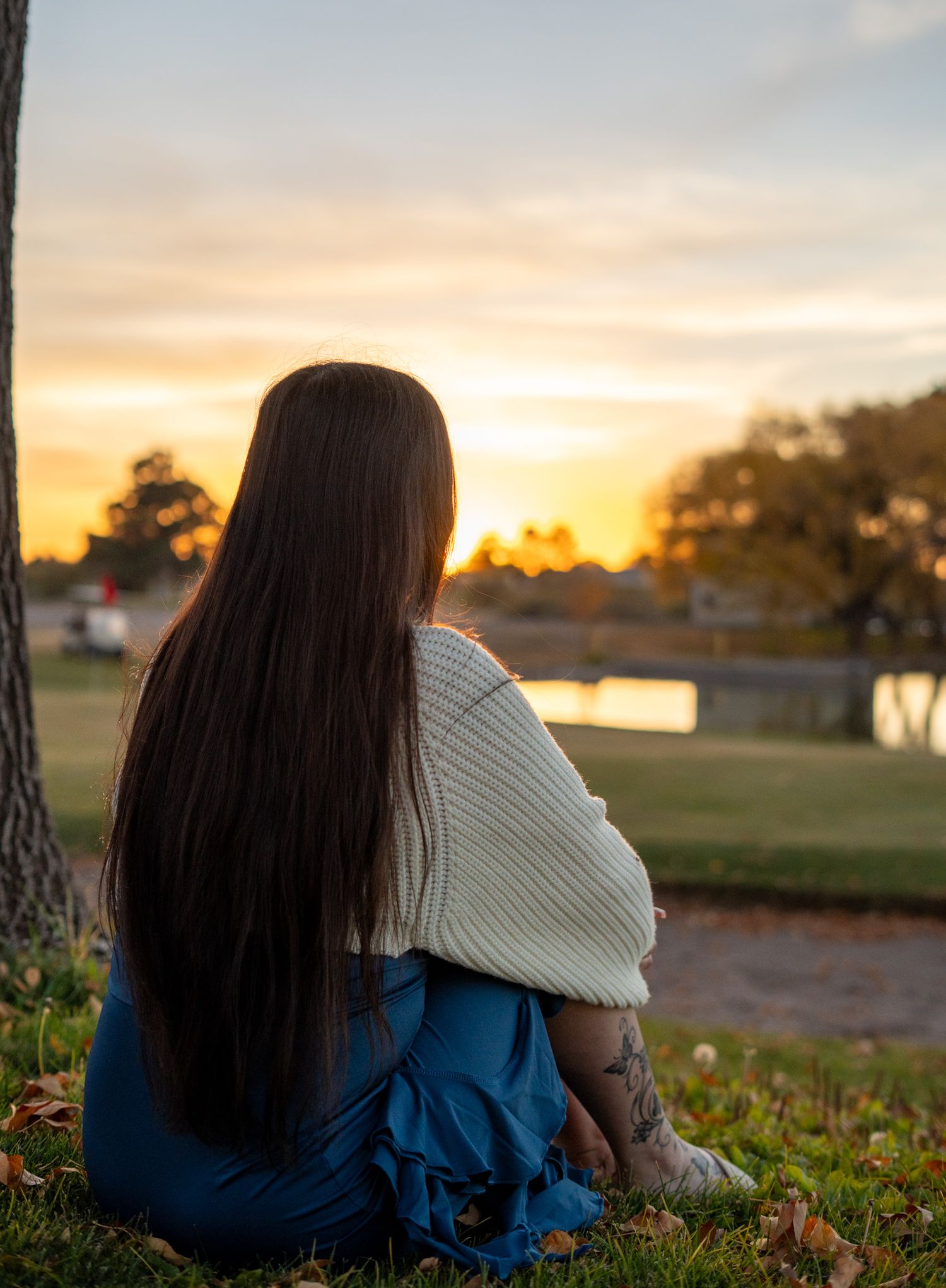 Person in cozy sweater sitting on grass watching sunset by lake with trees.