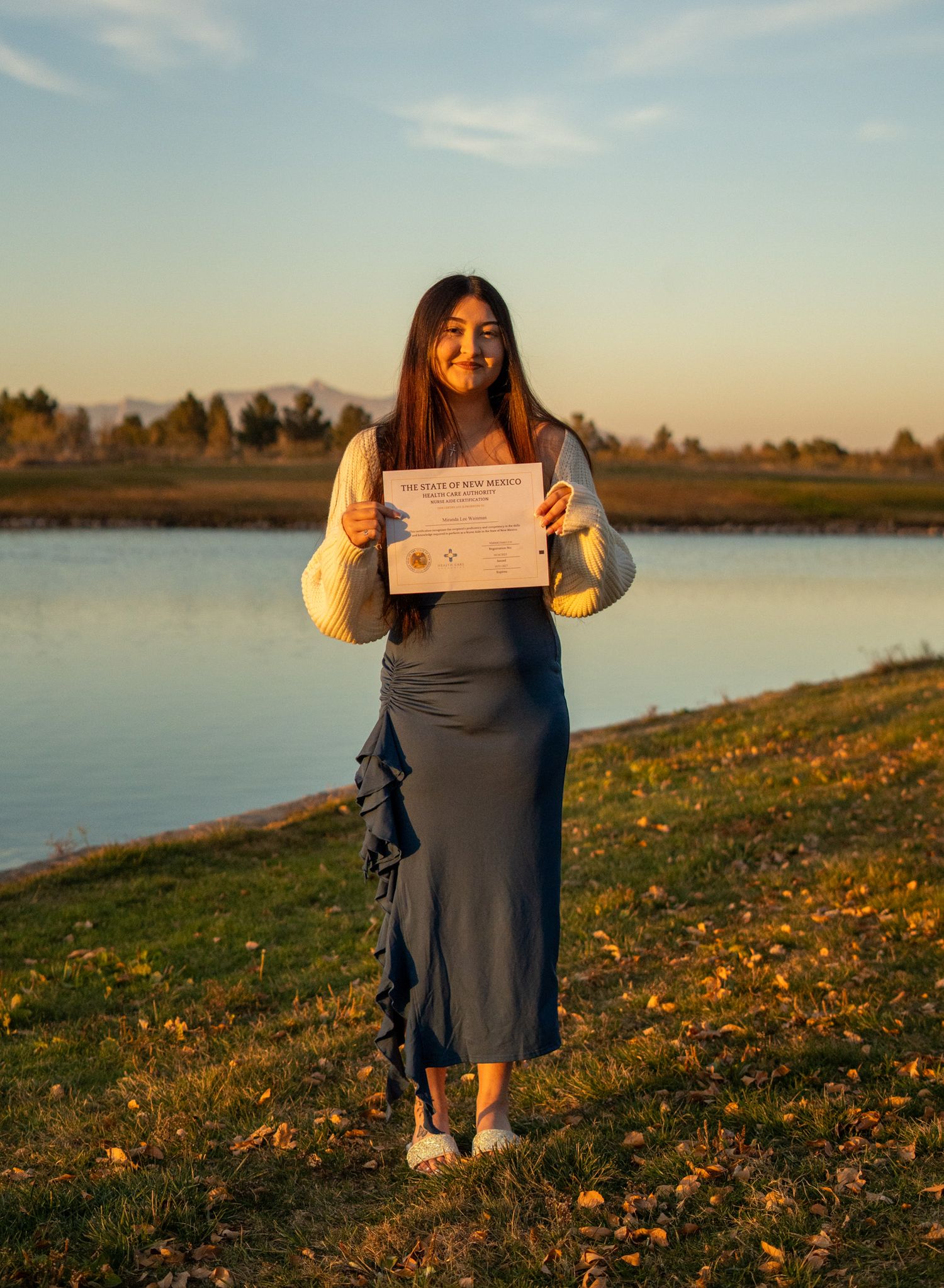 Person in blue dress poses with document on grassy lakeside during golden hour.