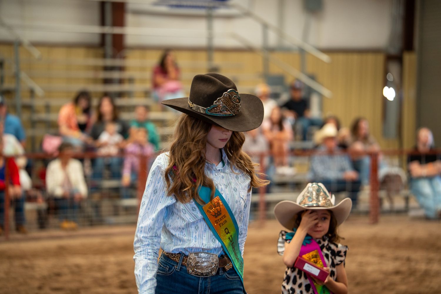 Western riders compete in a rodeo event inside a covered arena with spectators watching from the stands.
