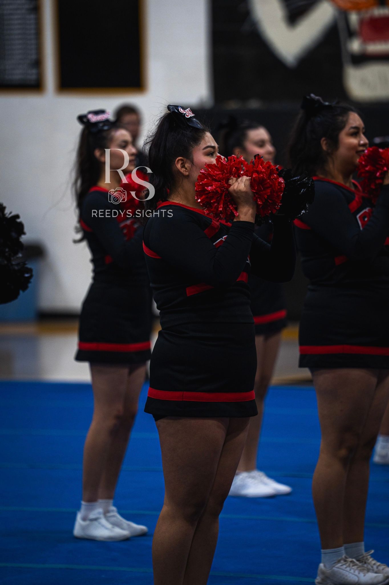 Cheerleaders in black uniforms with red pom poms perform a synchronized routine on a blue mat.