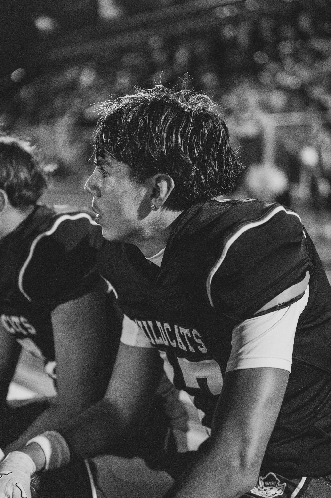 Black and white photo of a high school football player in uniform sitting on sidelines during a game.
