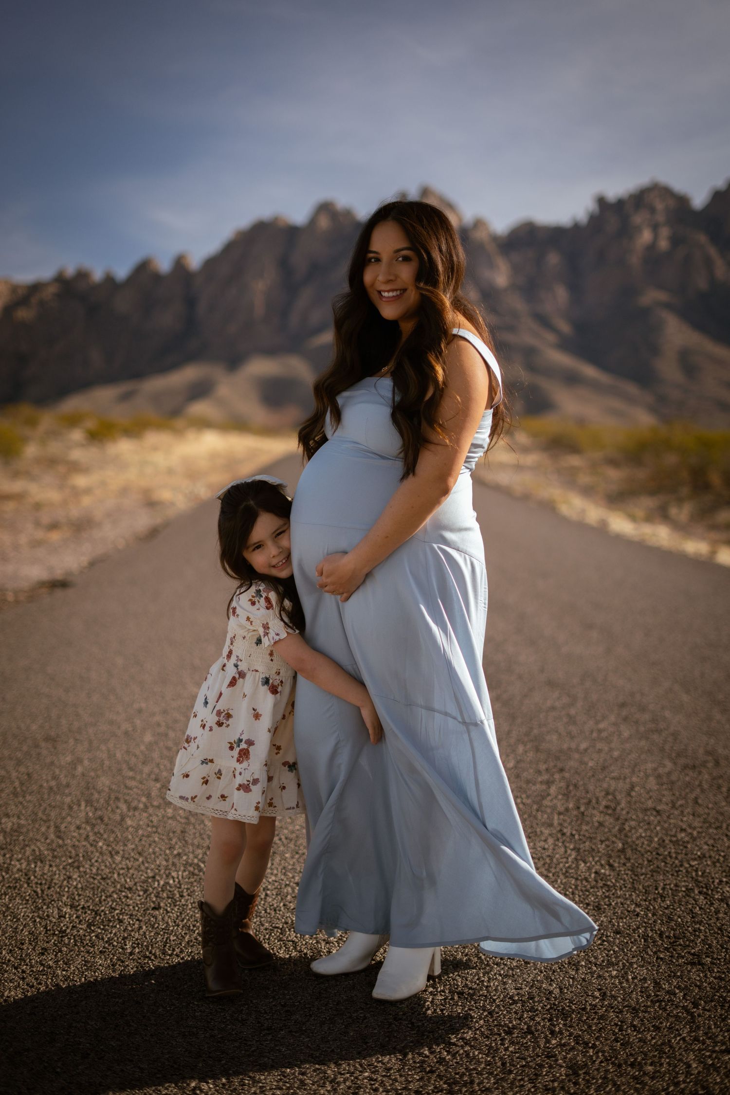 A touching maternity moment captured on a desert road with dramatic mountain peaks in the background.