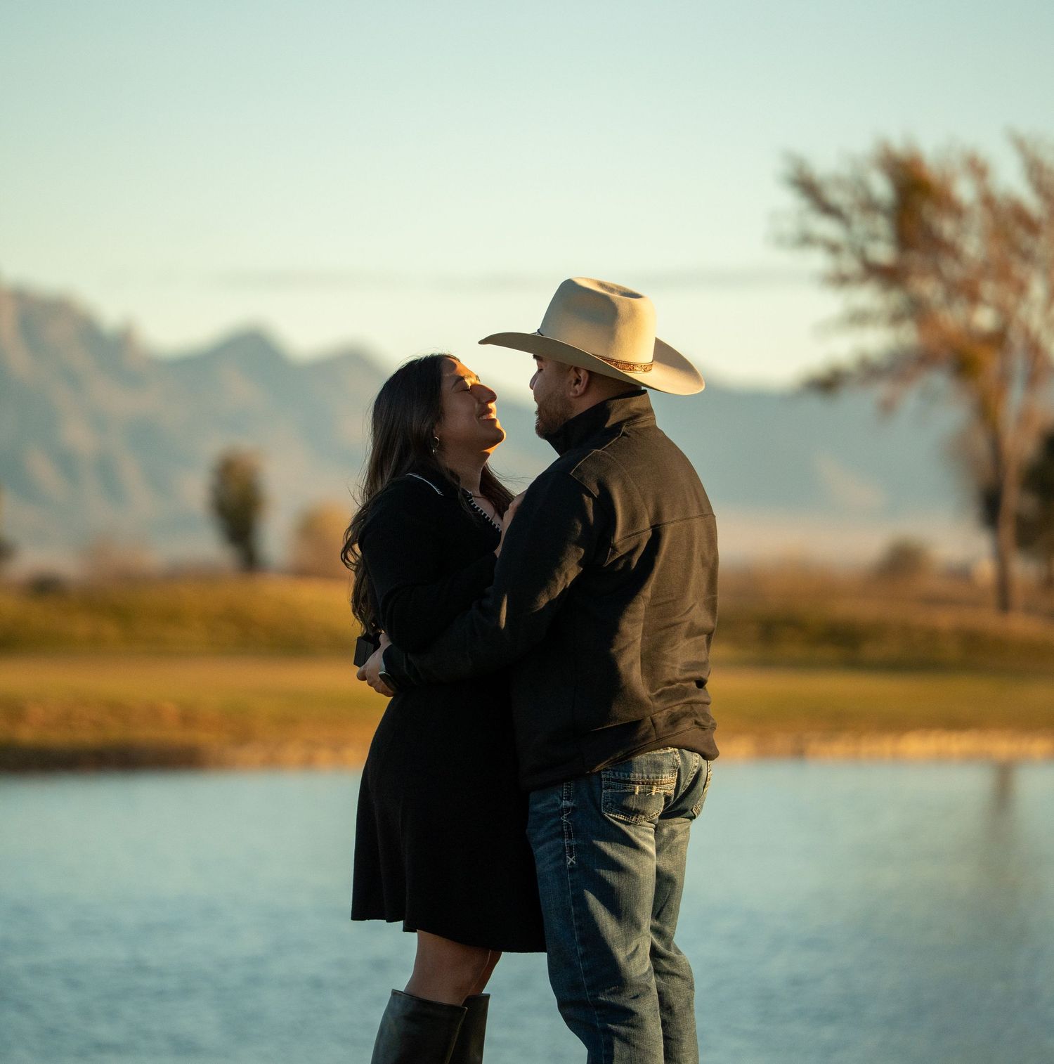 Silhouetted couple in cowboy hats share a romantic moment by a lake at sunset with mountains in the background.