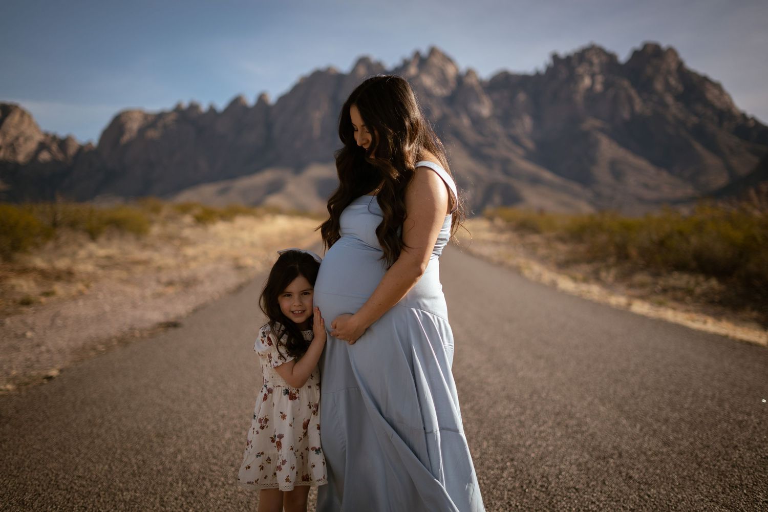 A mother and daughter share tender moments while walking together on a desert road at sunset.