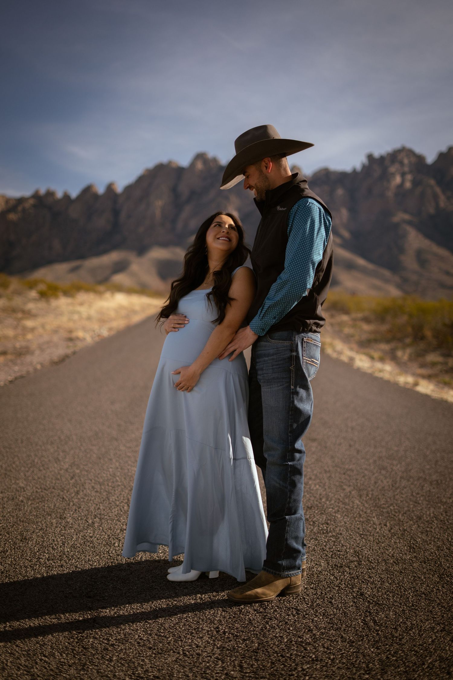 Couple in romantic poses on a desert road with mountains in the background at sunset.
