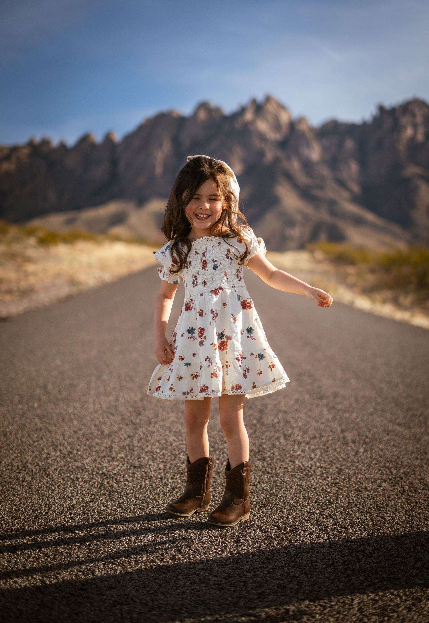A child in a floral dress and boots dances on a desert road with rugged mountains in the background.