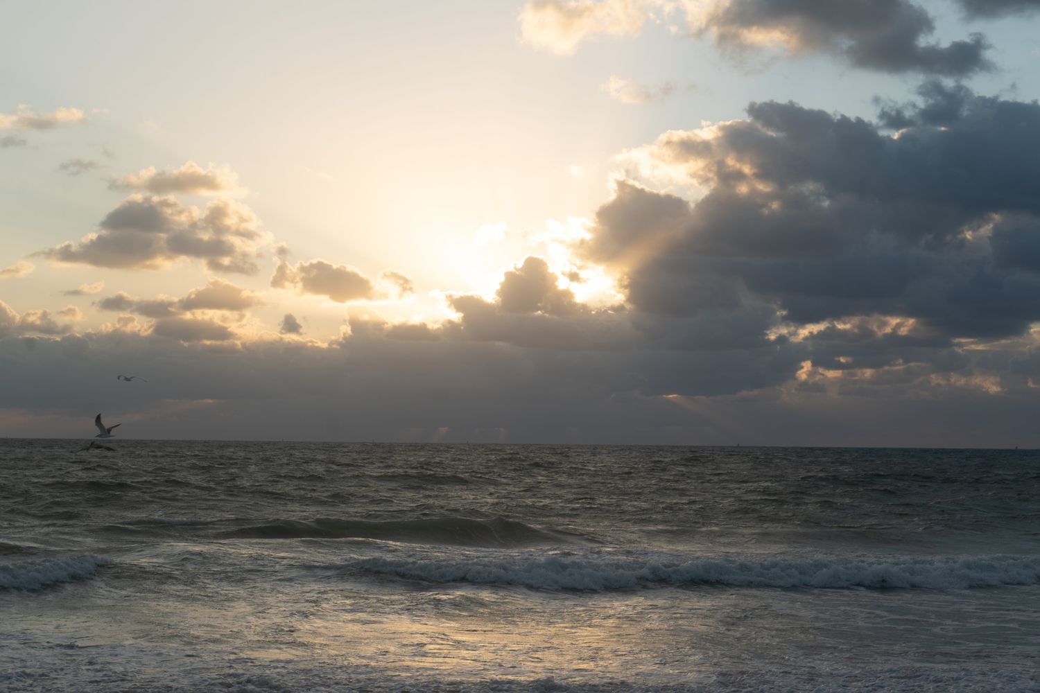 Dramatic sunset over ocean waves with sailboat silhouette on horizon.