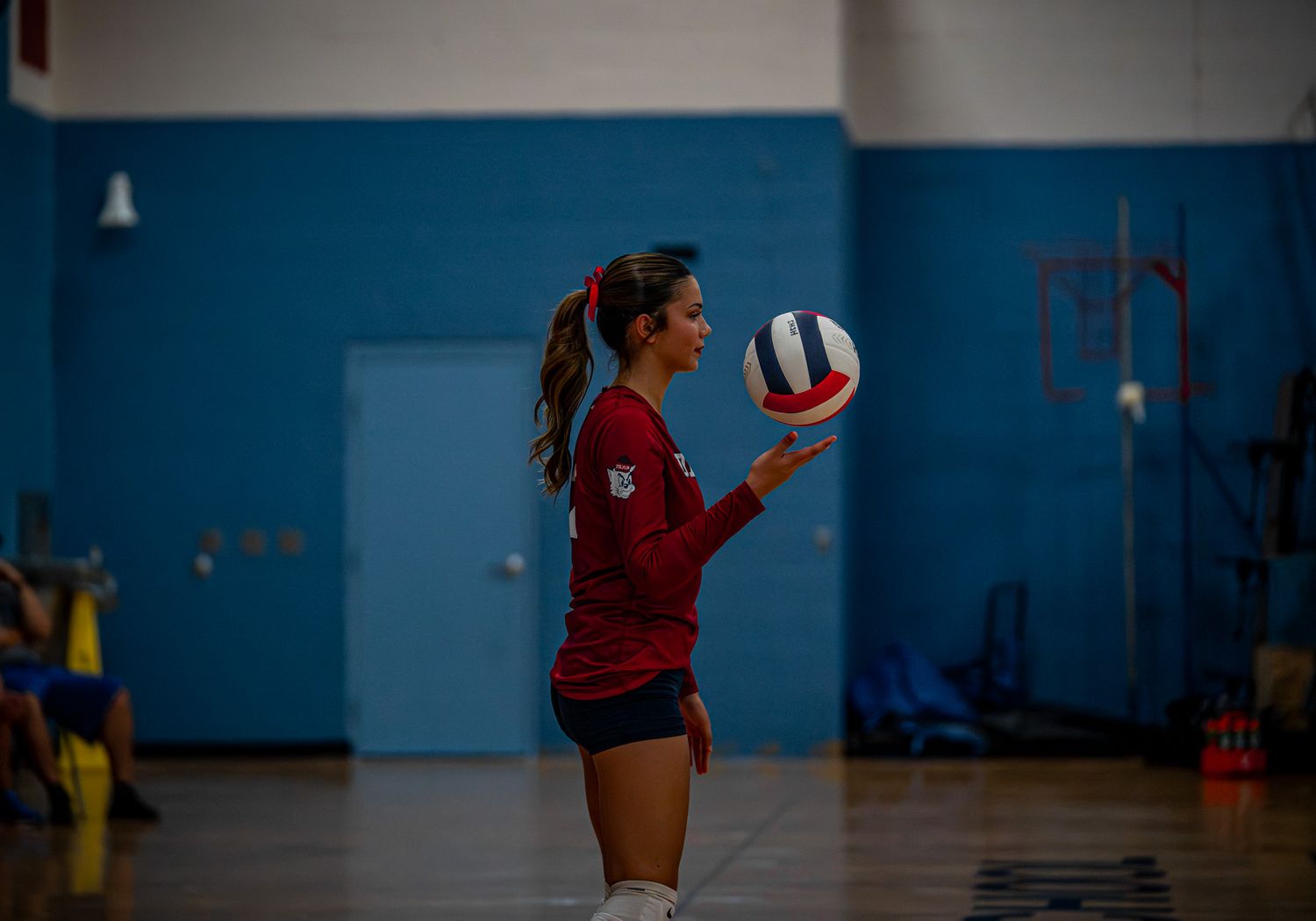 Volleyball player in red jersey preparing to serve in blue gymnasium.