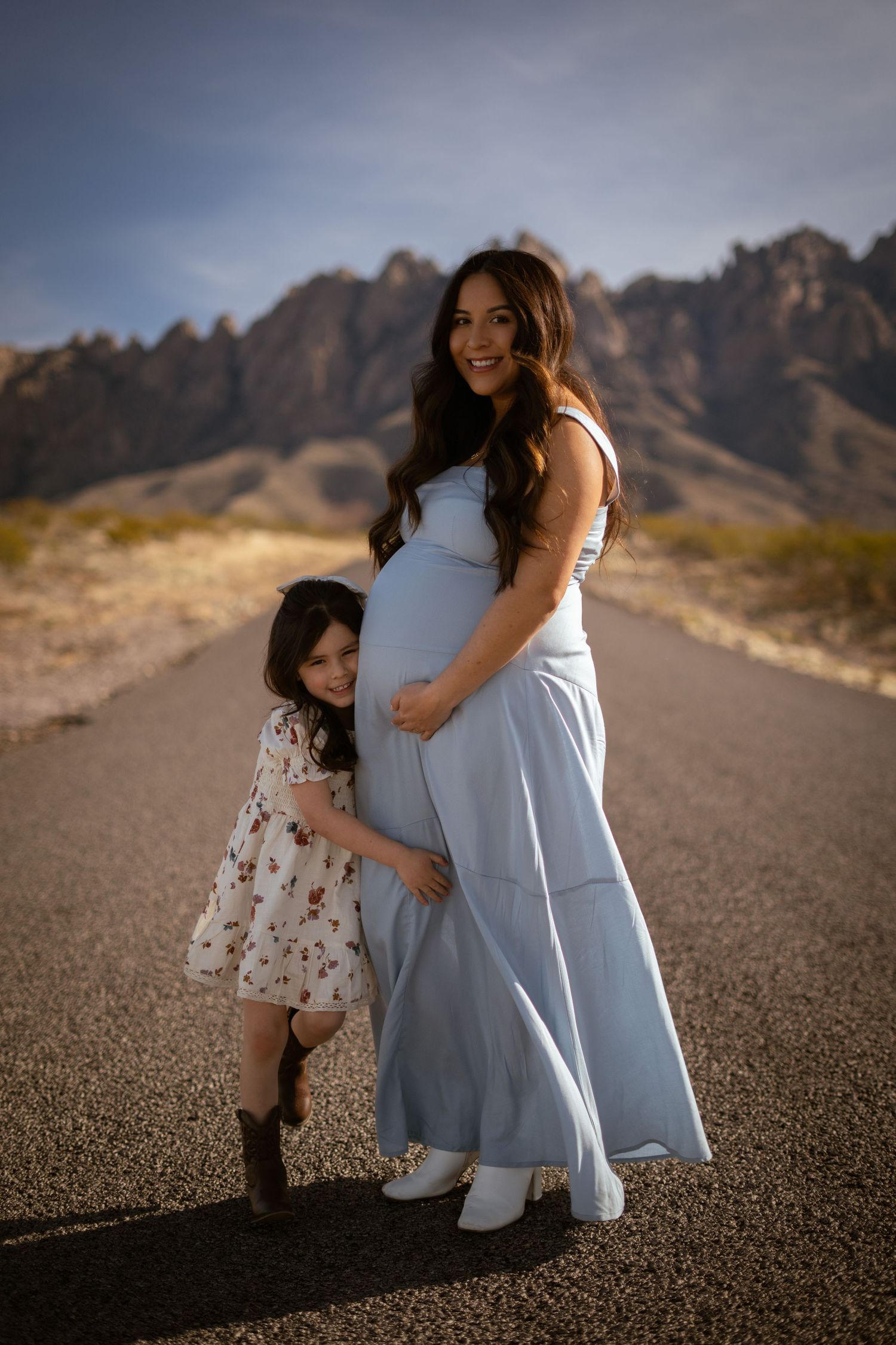 Mother and daughter share tender moments on desert road with rugged mountain backdrop at sunset.