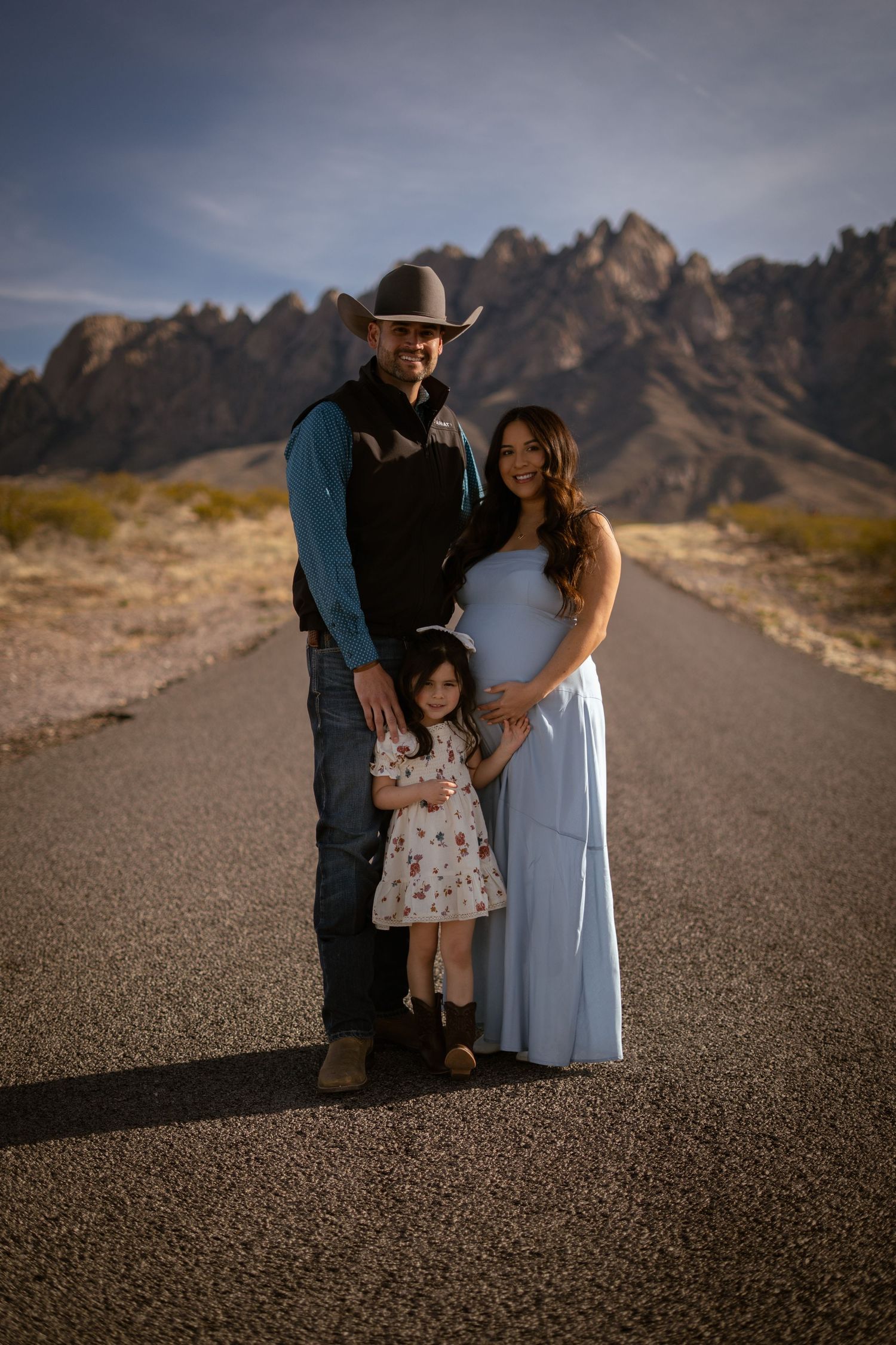 A family stands together on a desert road with dramatic mountain peaks in the background at sunset.