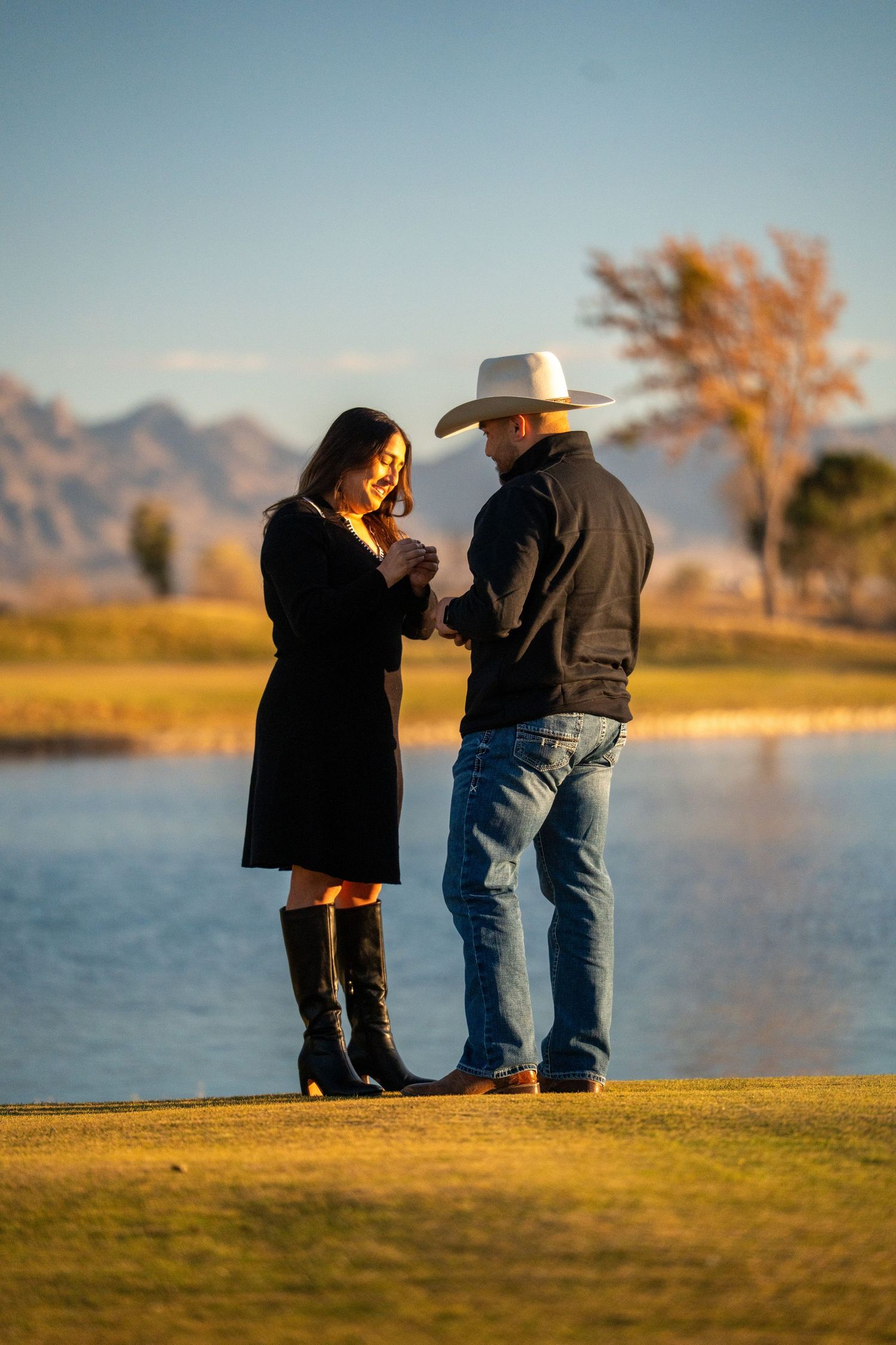 Couple standing together on grassy hill overlooking lake and mountains during golden hour.