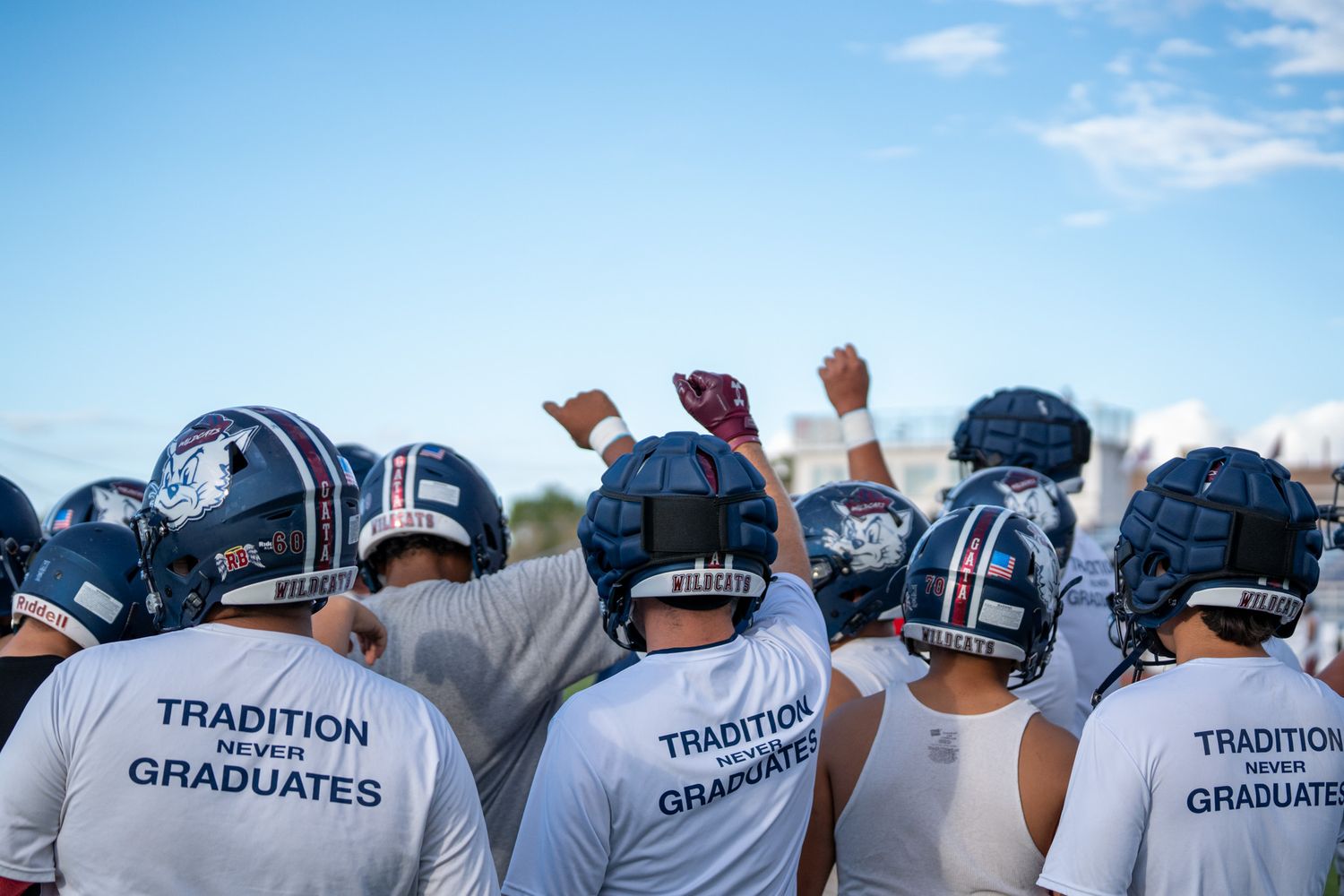 A group of people wearing matching white t-shirts with 'TRADITION GRADUATES' text stand together outdoors under blue sky.