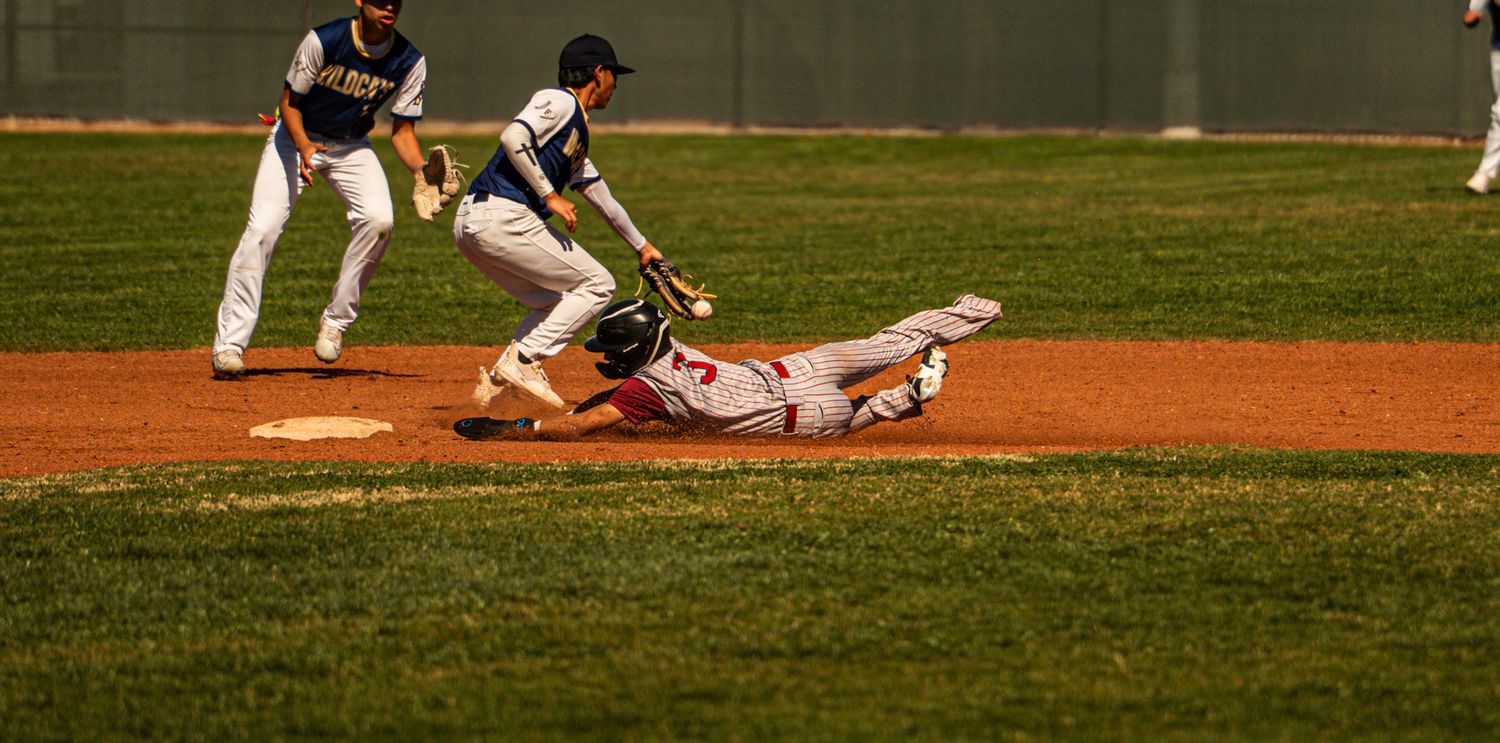 A baseball player in a dark uniform swings the bat during a game on a sunny day at the baseball field.