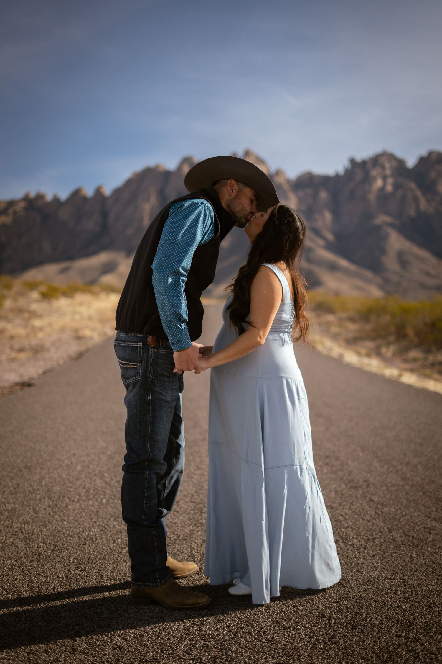 Two figures share a tender moment on a desert road with rugged mountains silhouetted against the sky.
