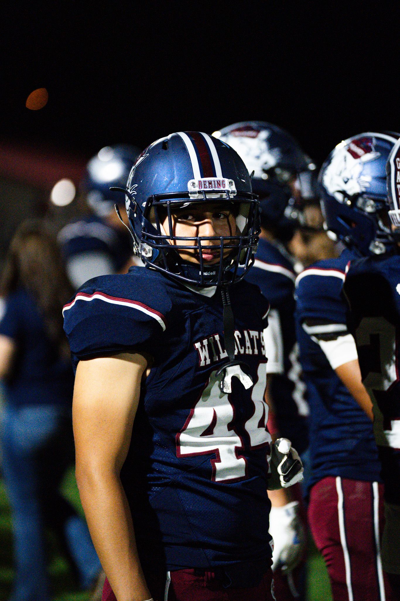 Football team stands in formation wearing navy blue uniforms and helmets during a night game.