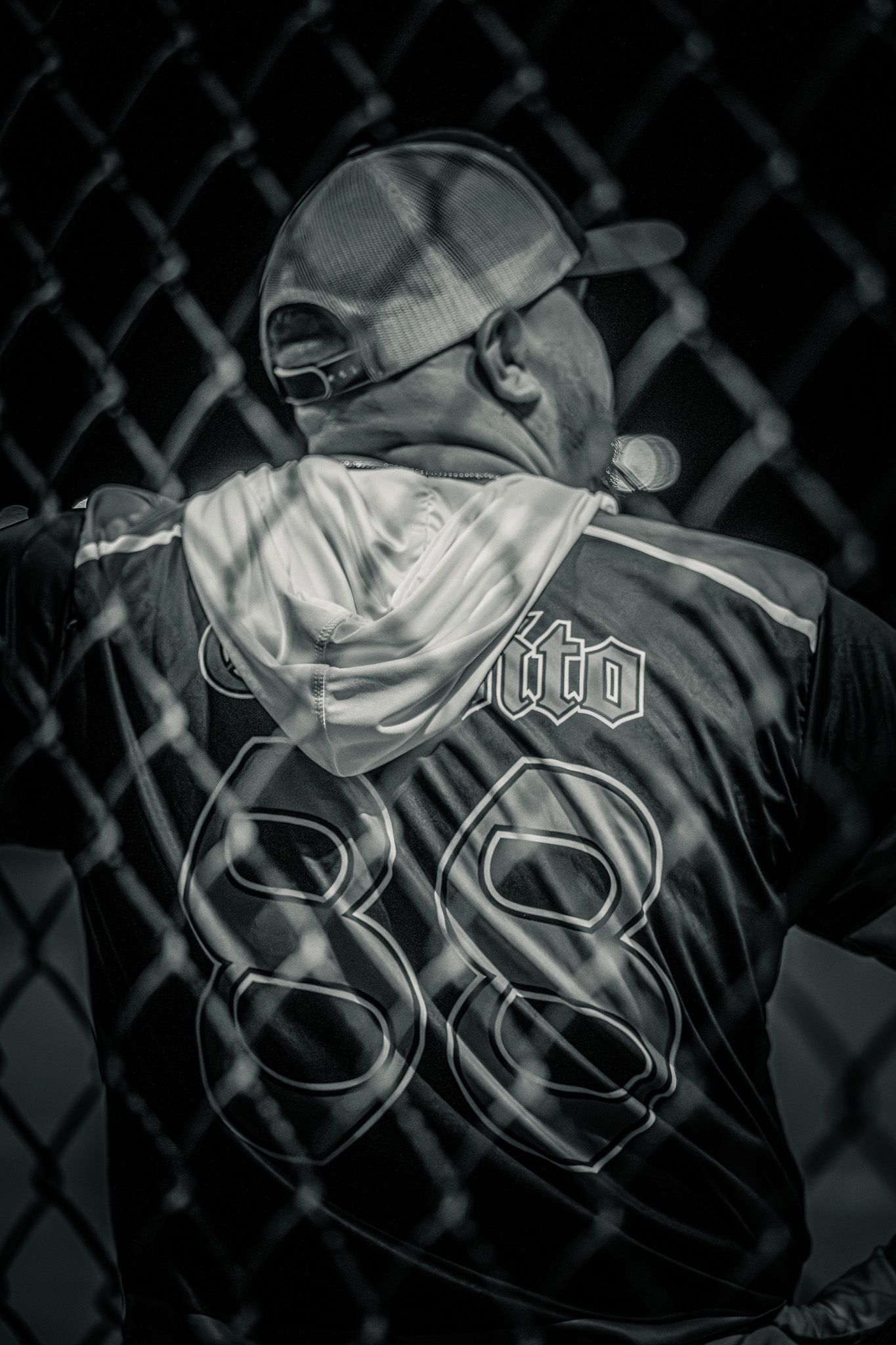 A black and white image shows someone wearing a baseball cap with chain link fence in the background.