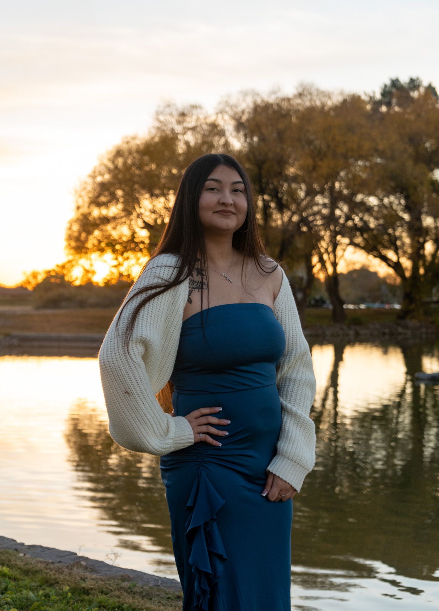 A woman in a blue dress and cream cardigan poses by a lake at sunset.