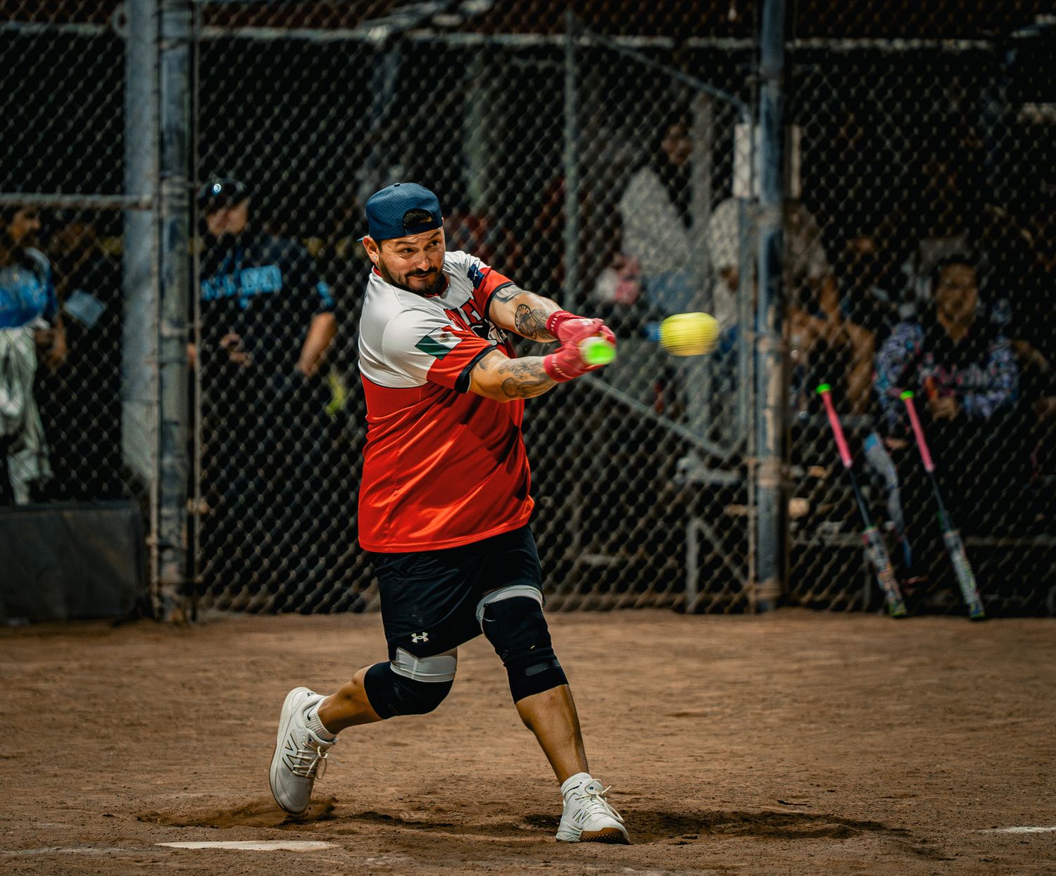 A softball player in a red and black uniform takes a powerful swing at an incoming pitch on a dirt field.