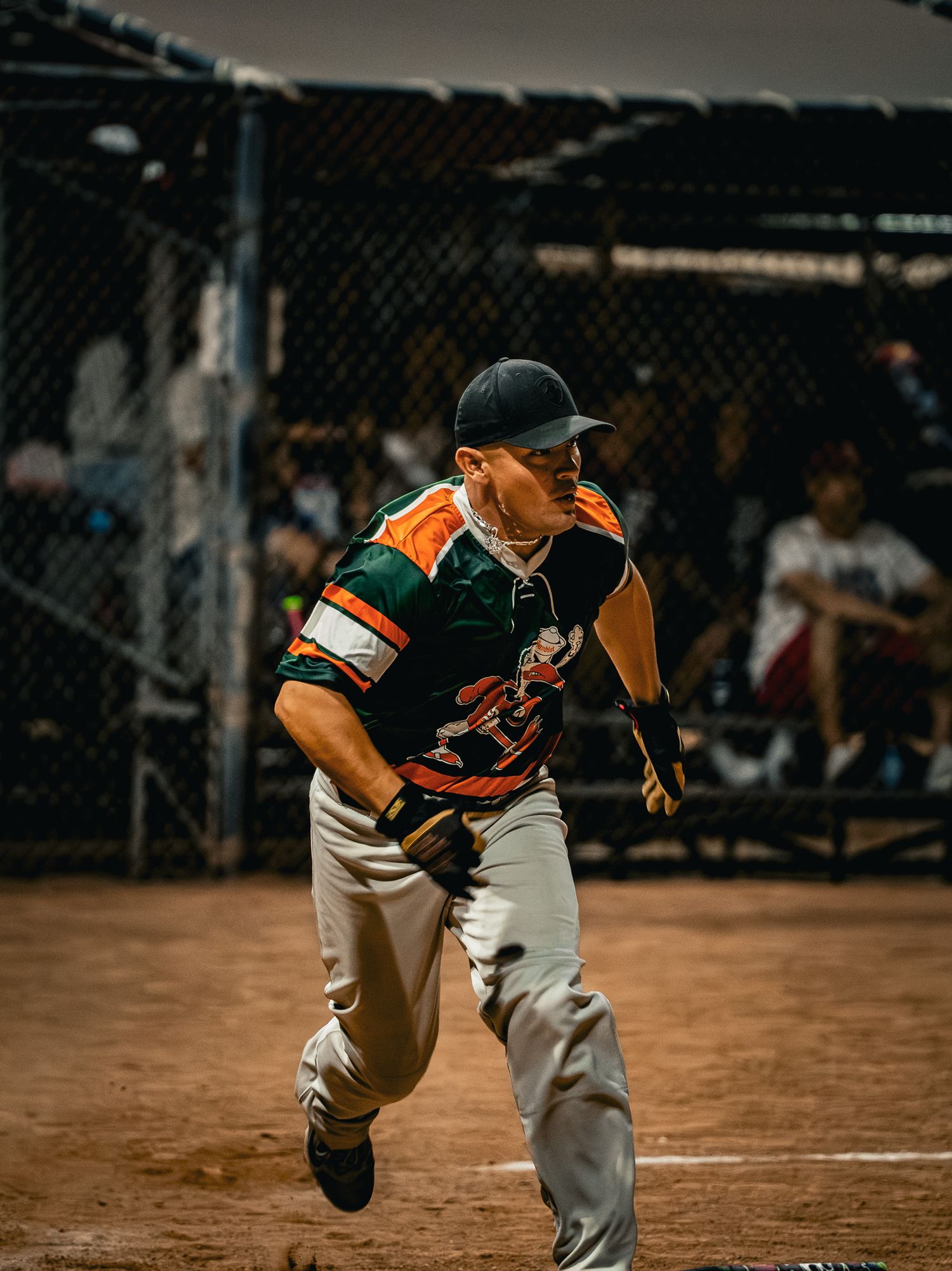 Baseball player in orange and green uniform slides into base during night game on dirt field.
