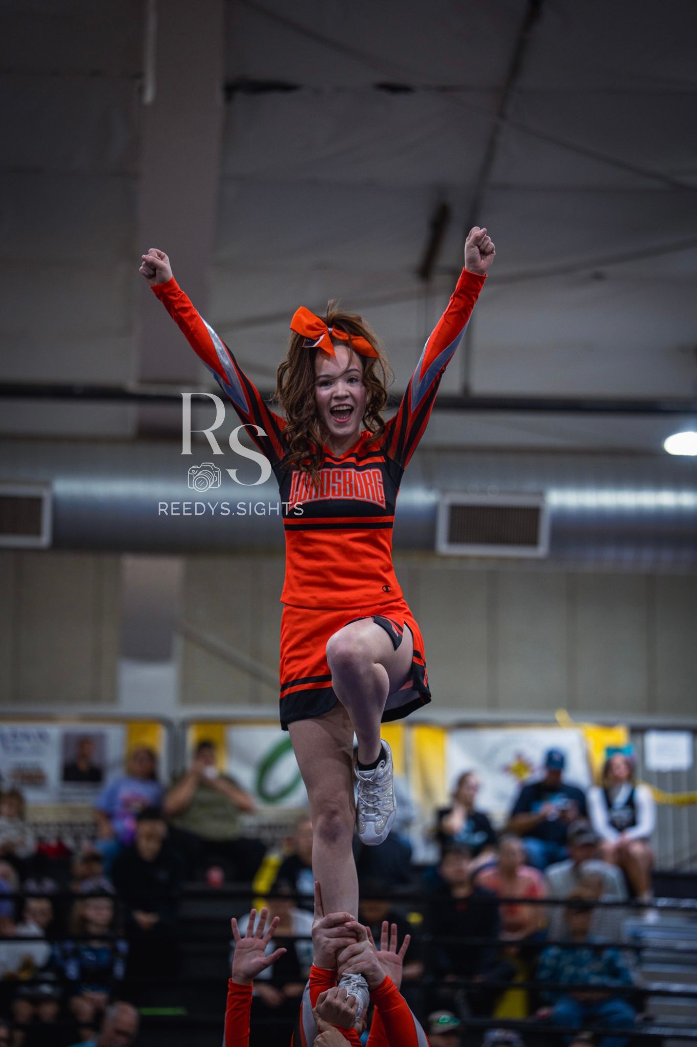 Cheerleader in orange uniform performs a high-flying stunt during indoor competition.
