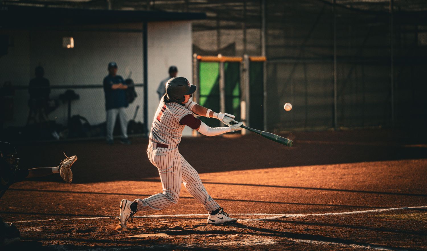 Baseball player in pinstripe uniform swings bat at night game on dirt field under stadium lights.