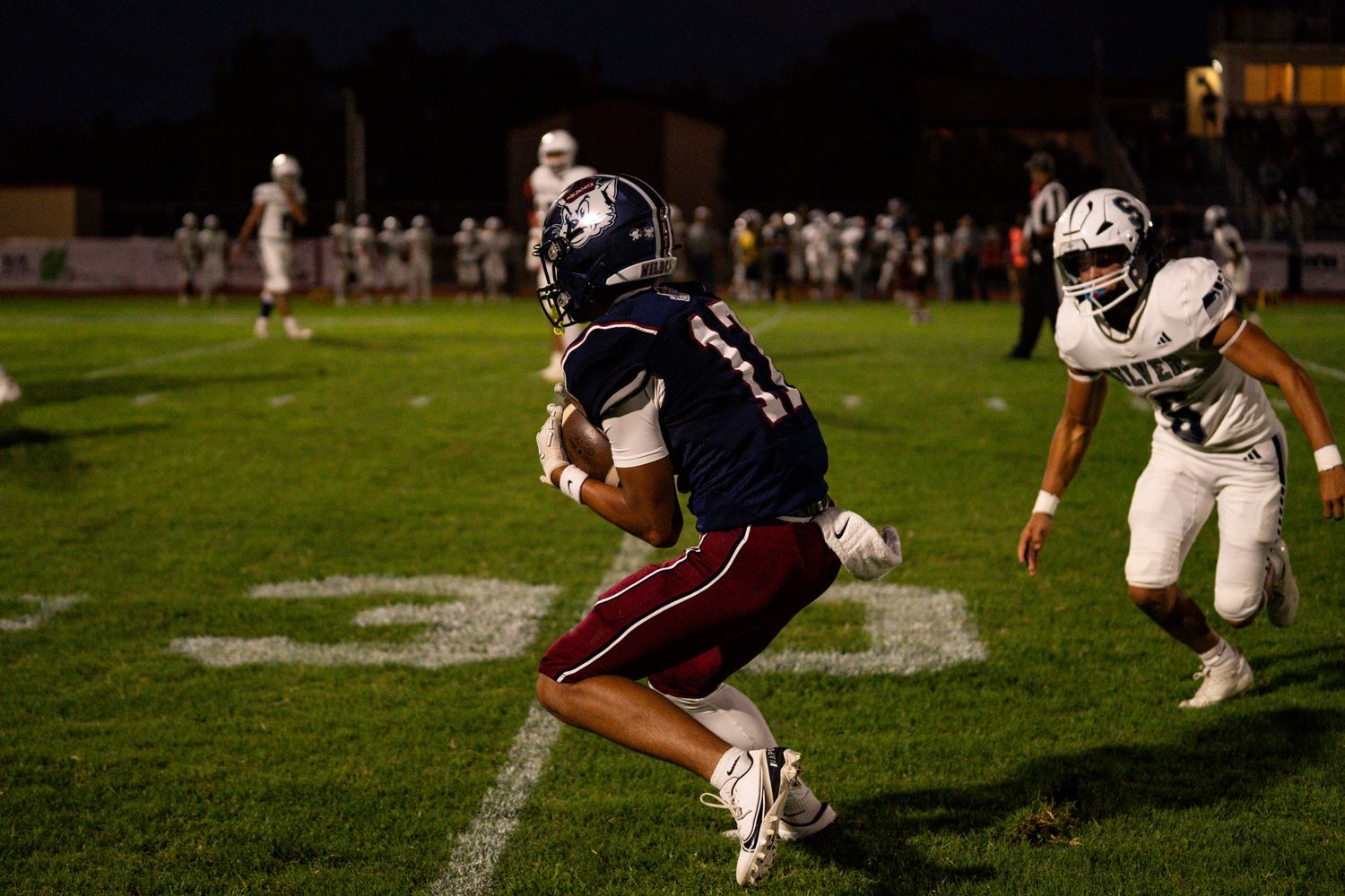 A football player in a dark uniform attempts a diving tackle during a nighttime game on a grass field.
