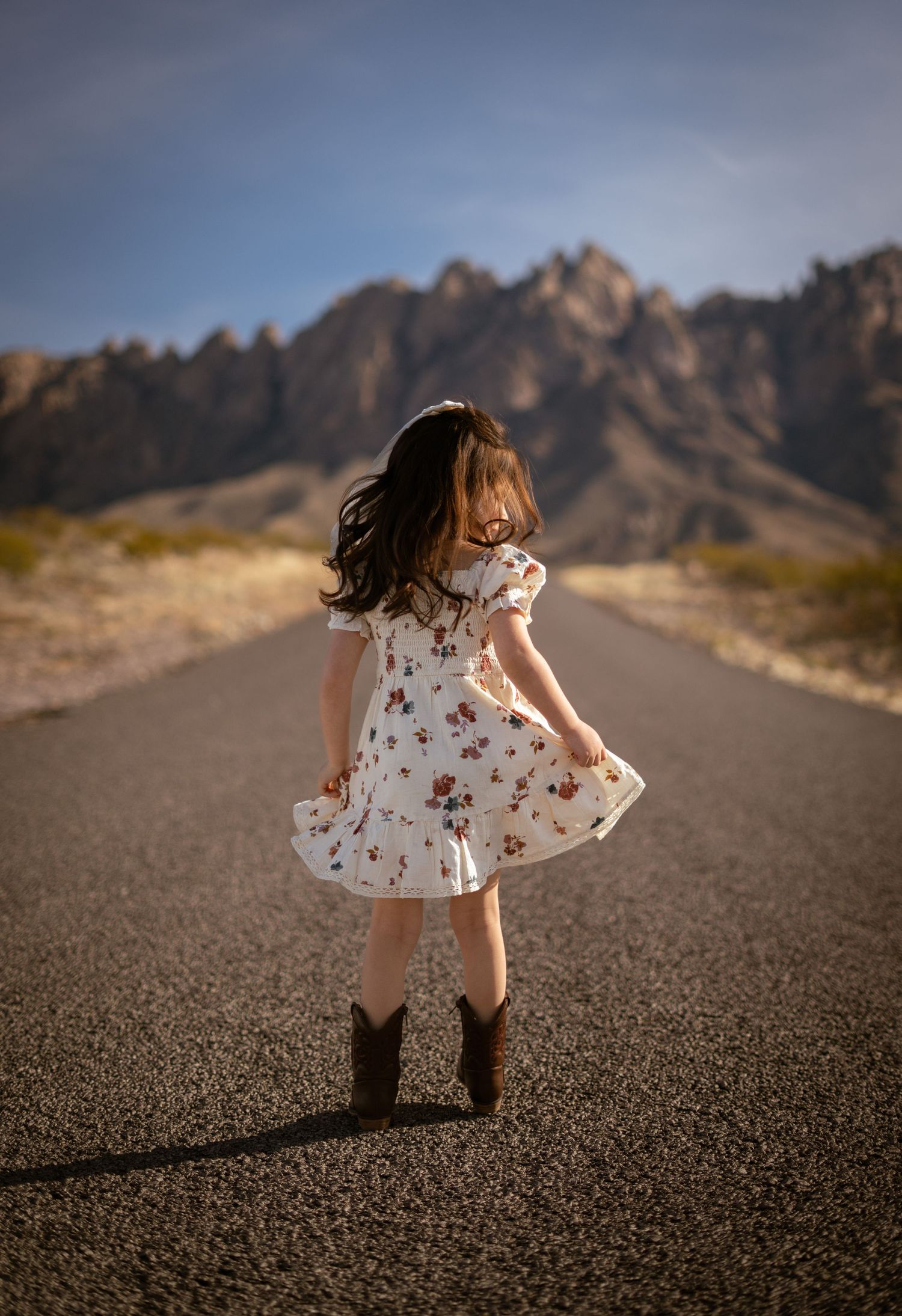 A young child in a floral dress explores a desert road with rugged mountain peaks in the background.