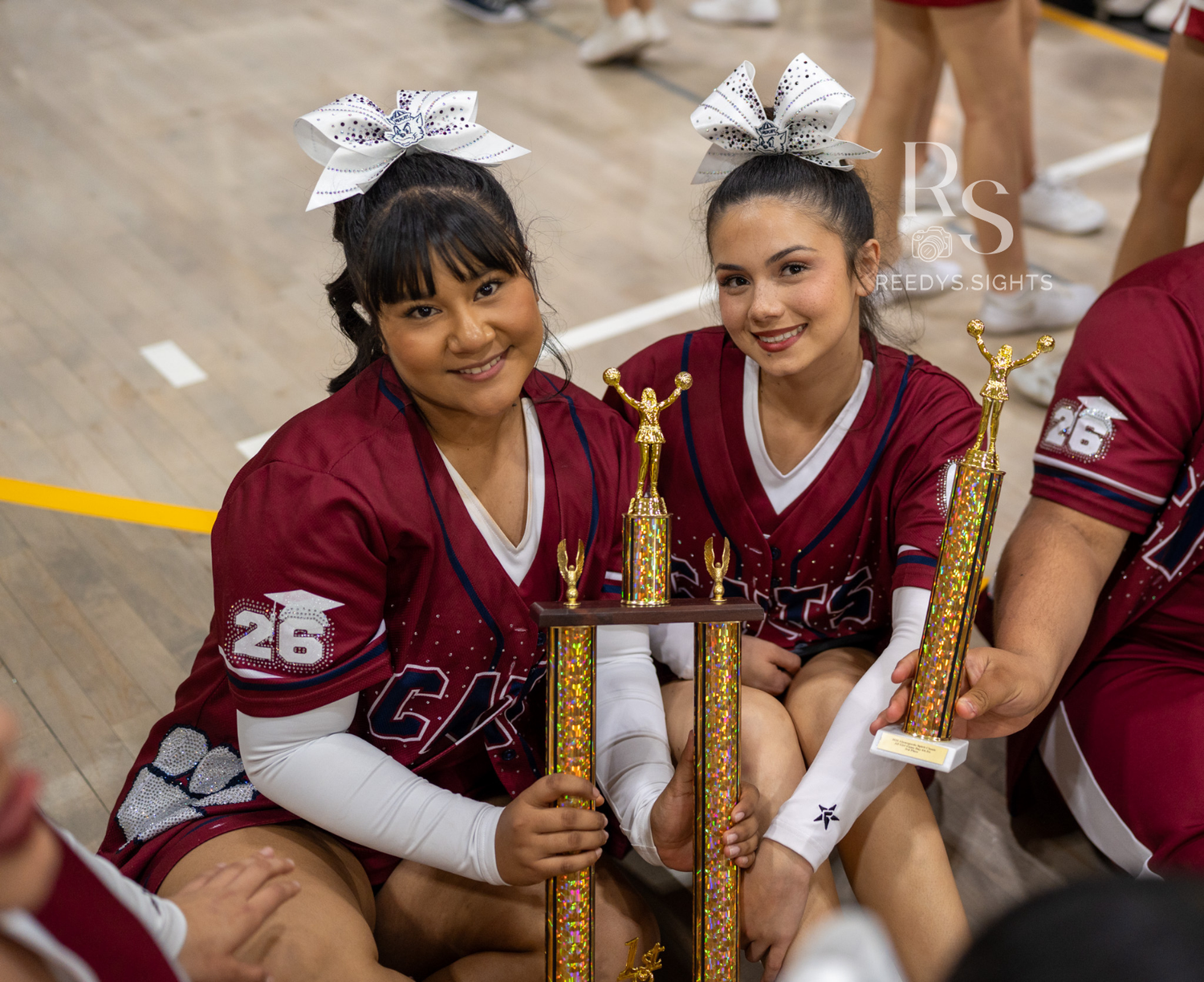 Youth softball teammates in maroon jerseys celebrate with their tournament trophies after a game.
