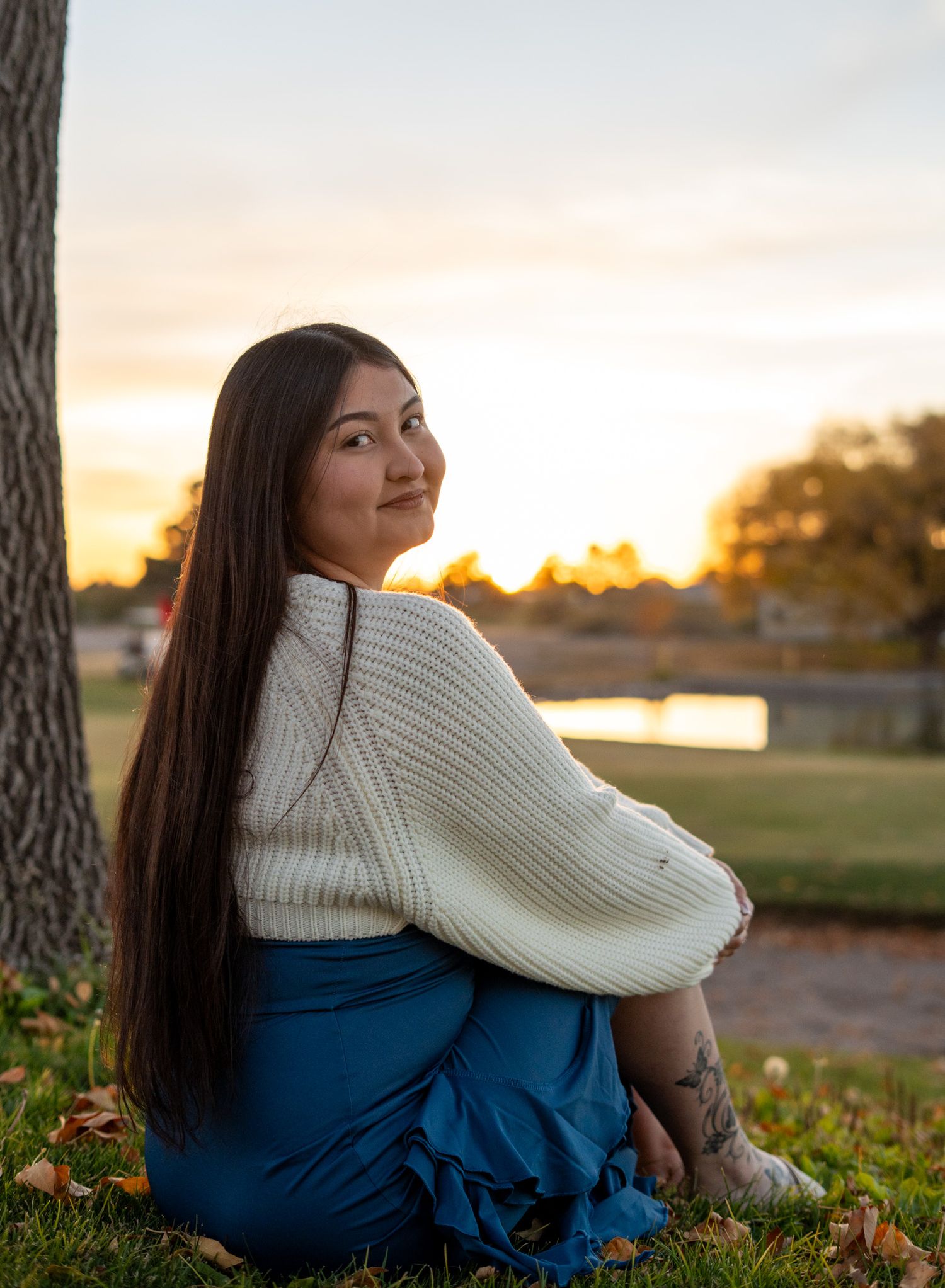 Person in white sweater and blue dress sits on grass against sunset backdrop.