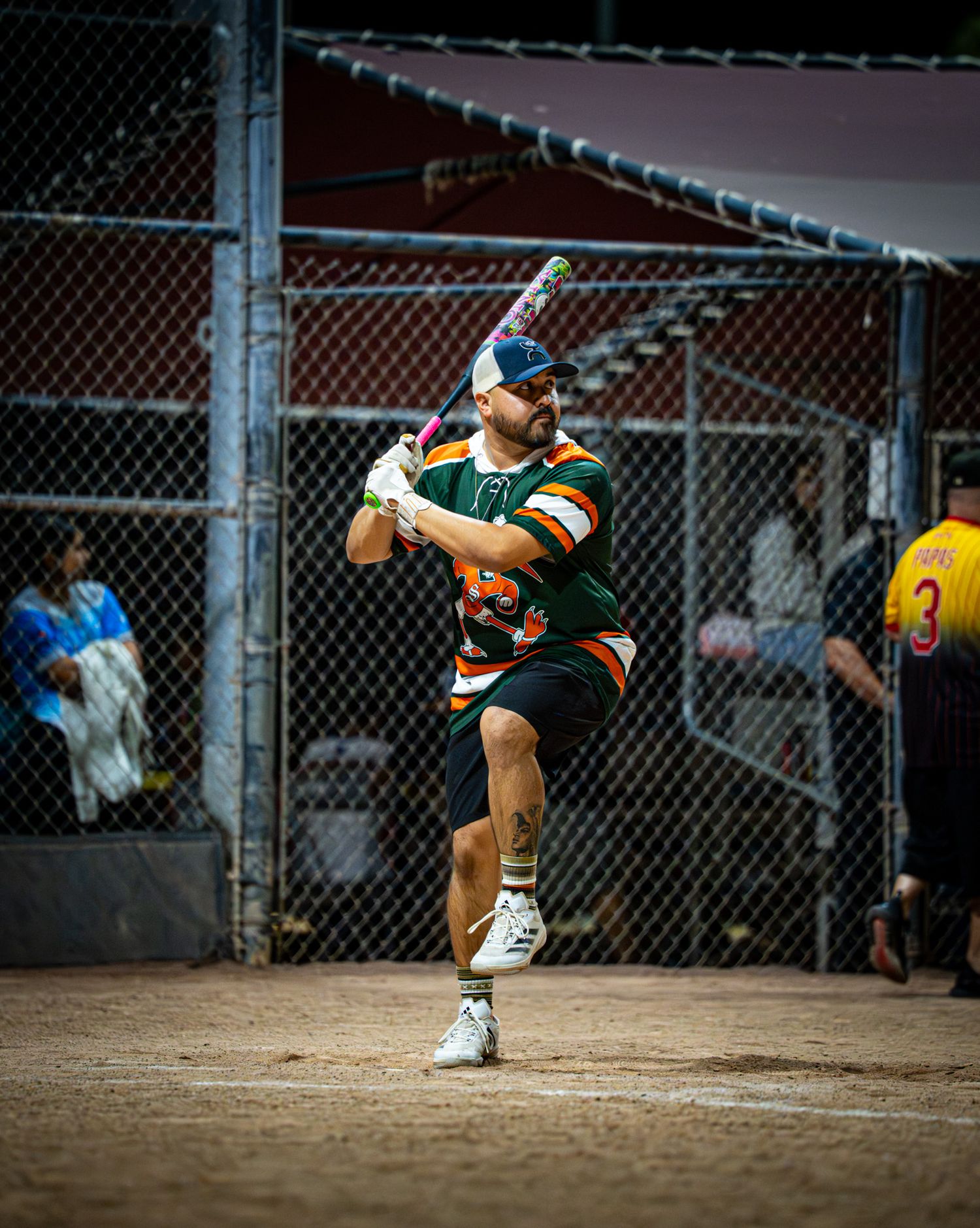 A softball player in a green and white uniform swings their bat during a competitive game at a fenced field.
