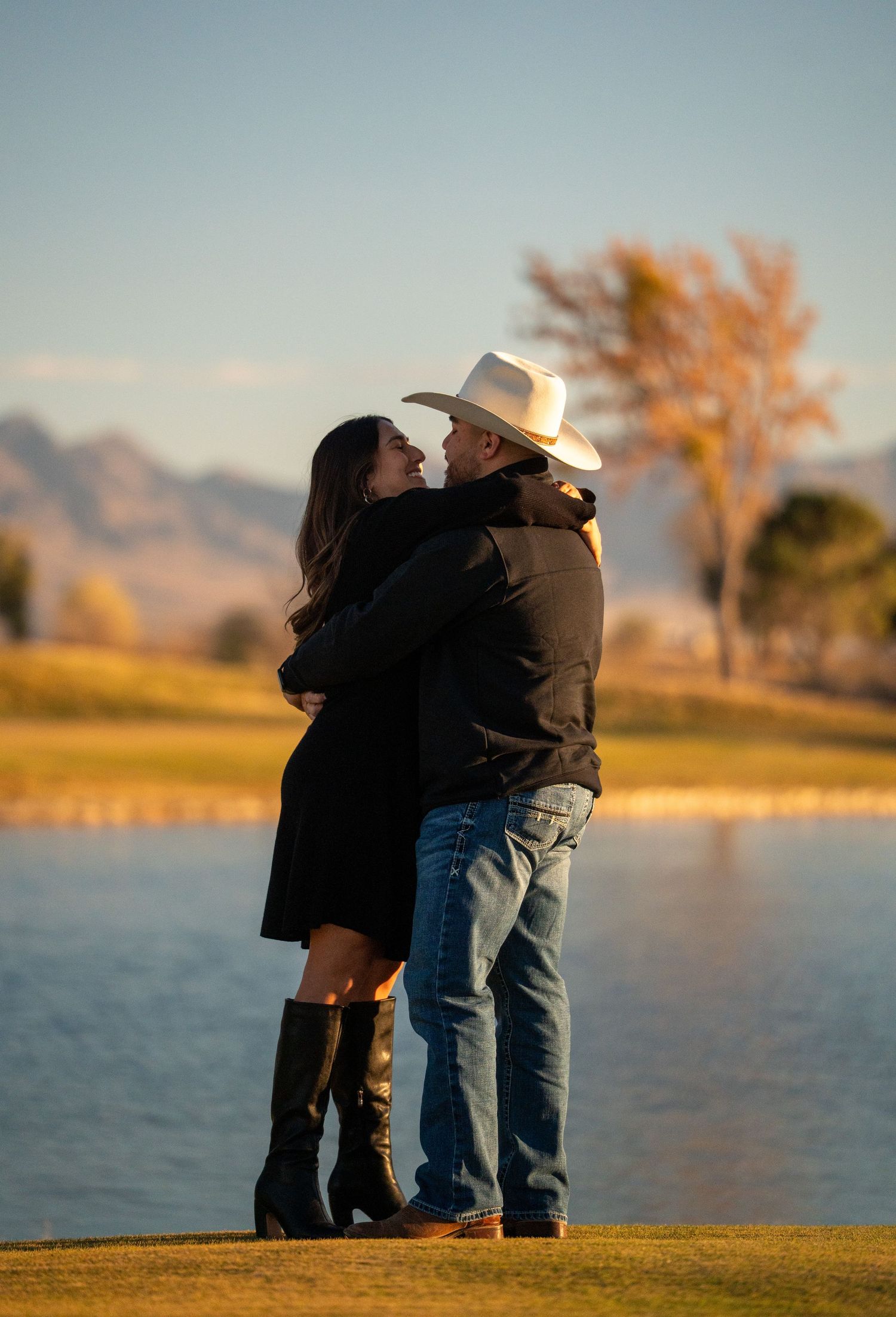 A couple stands together at sunset by a serene lake with autumn trees and mountains in the background.