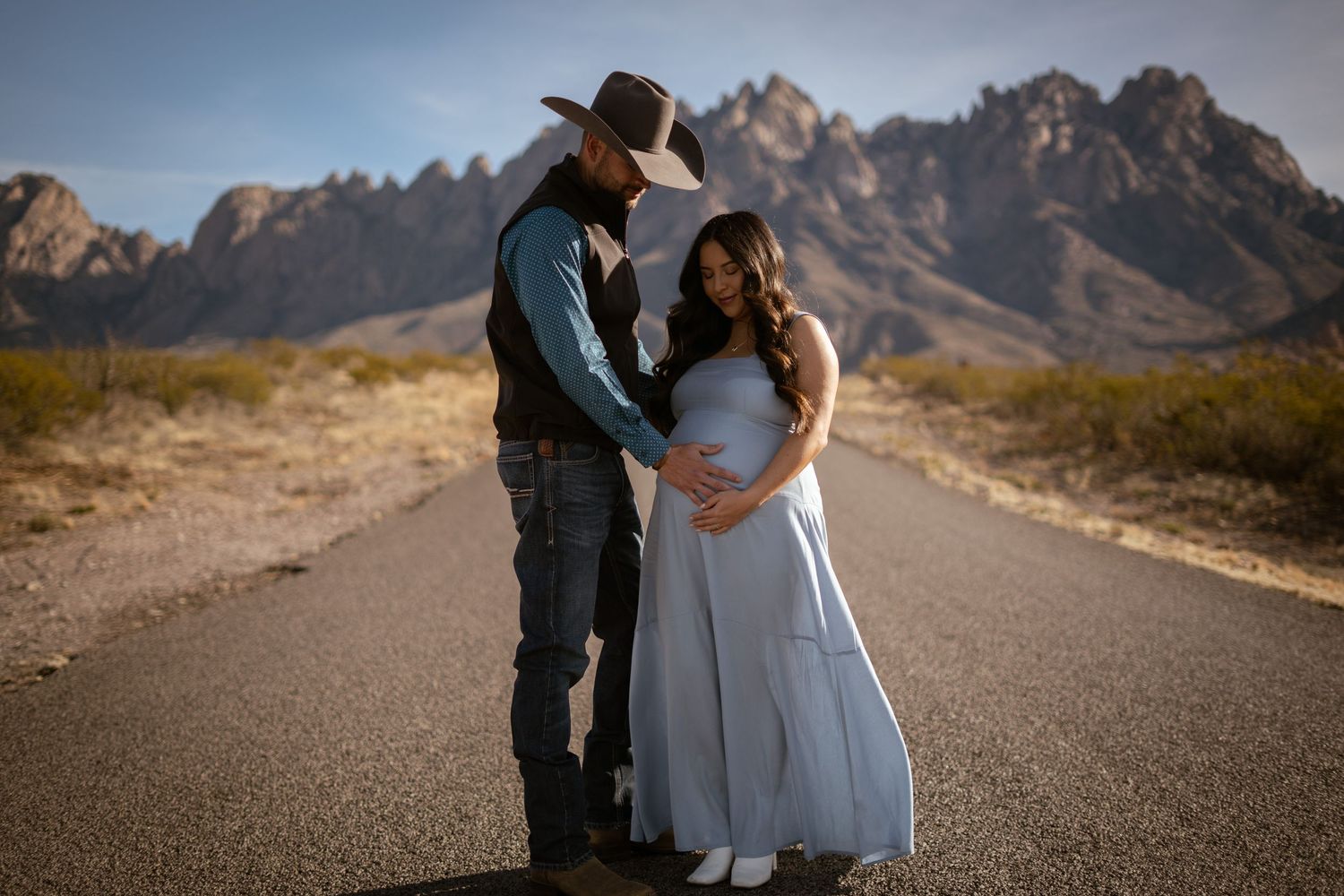 A couple shares a romantic moment on a desert road with rugged mountains in the background during golden hour.