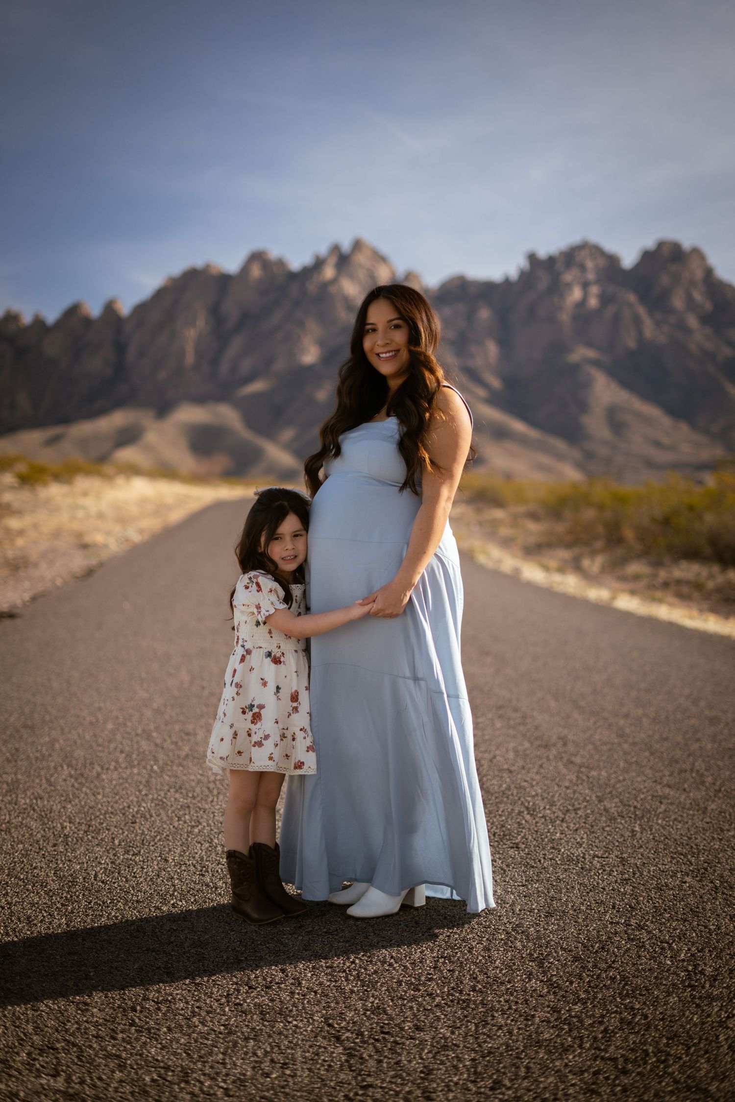 A tender moment captured on a desert road with mountains in the background as figures embrace in golden light.
