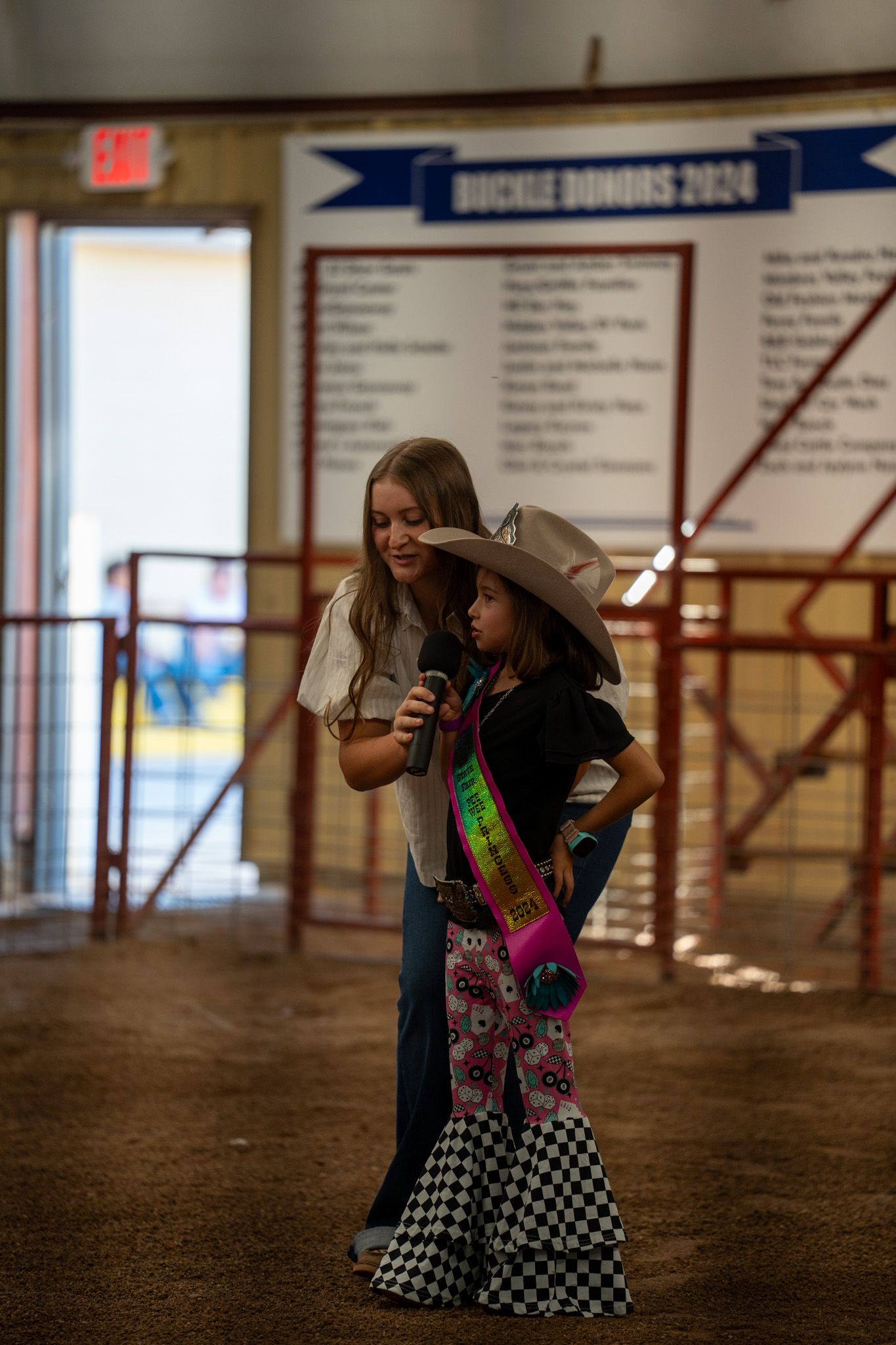 A young participant in western attire with a sash speaks into a microphone at an indoor rodeo event.