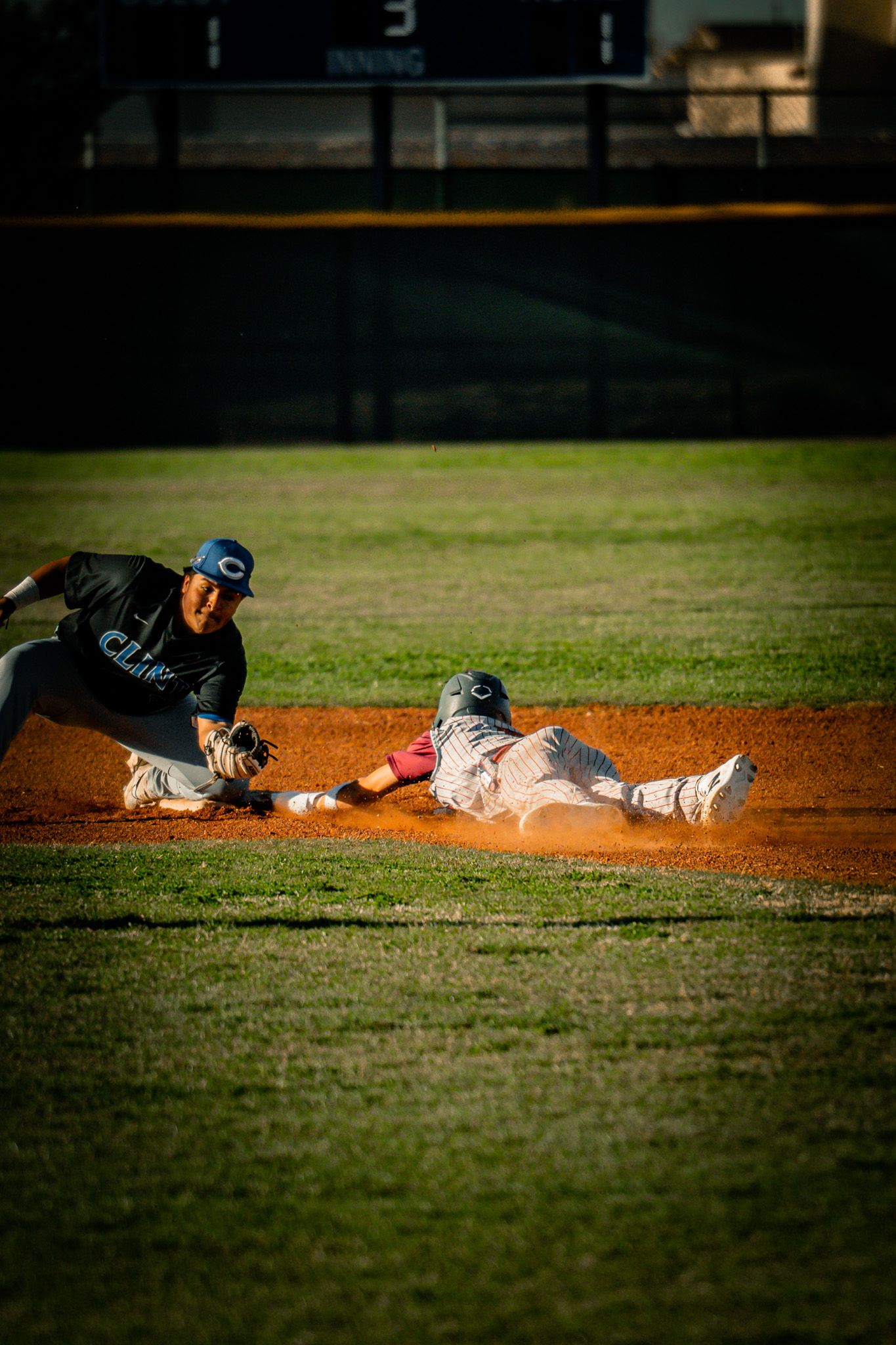Baseball players slide into bases during an intense game on a dirt infield at sunset.
