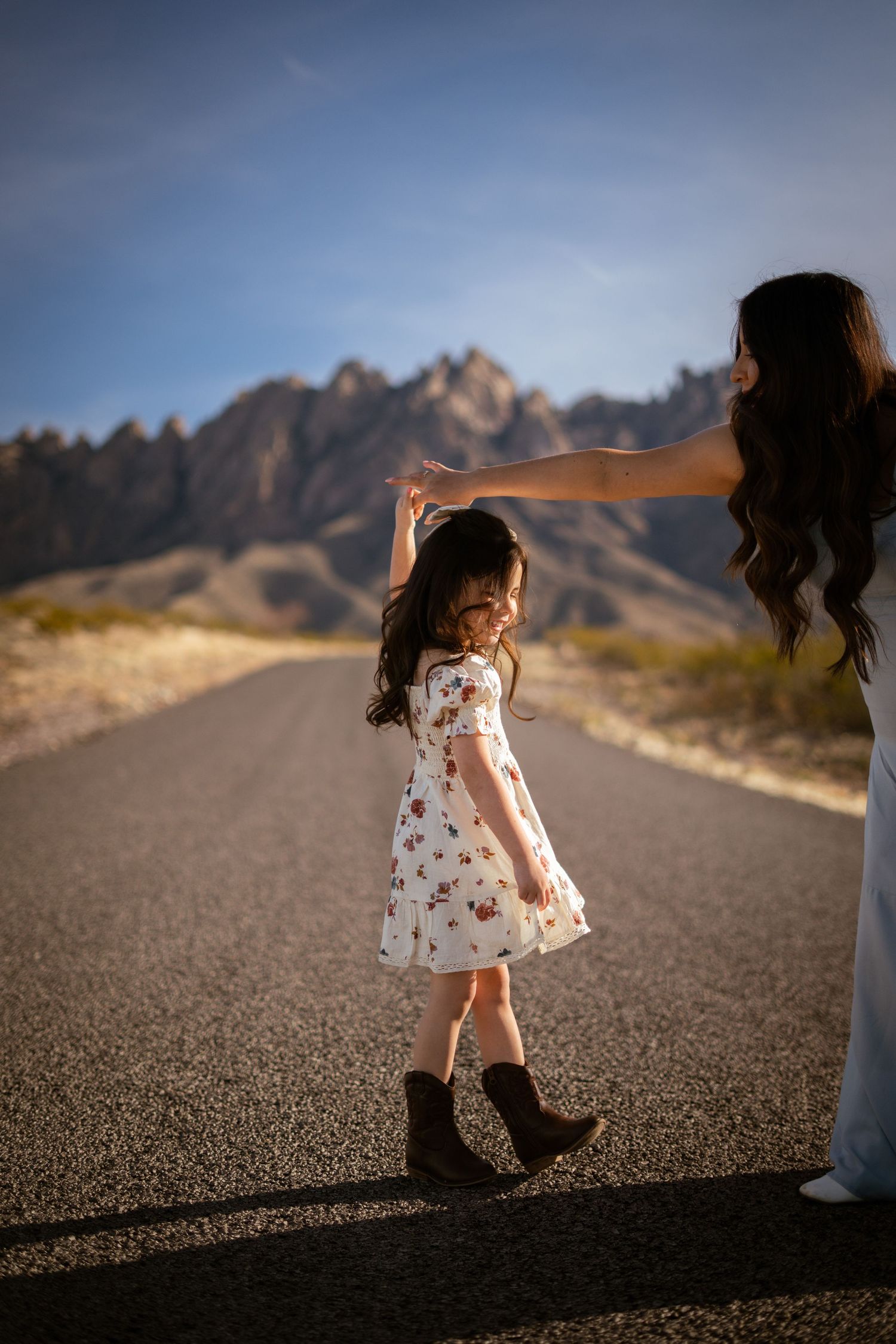 A small child in a white dress walks down an empty desert road with mountains in the background at sunset.