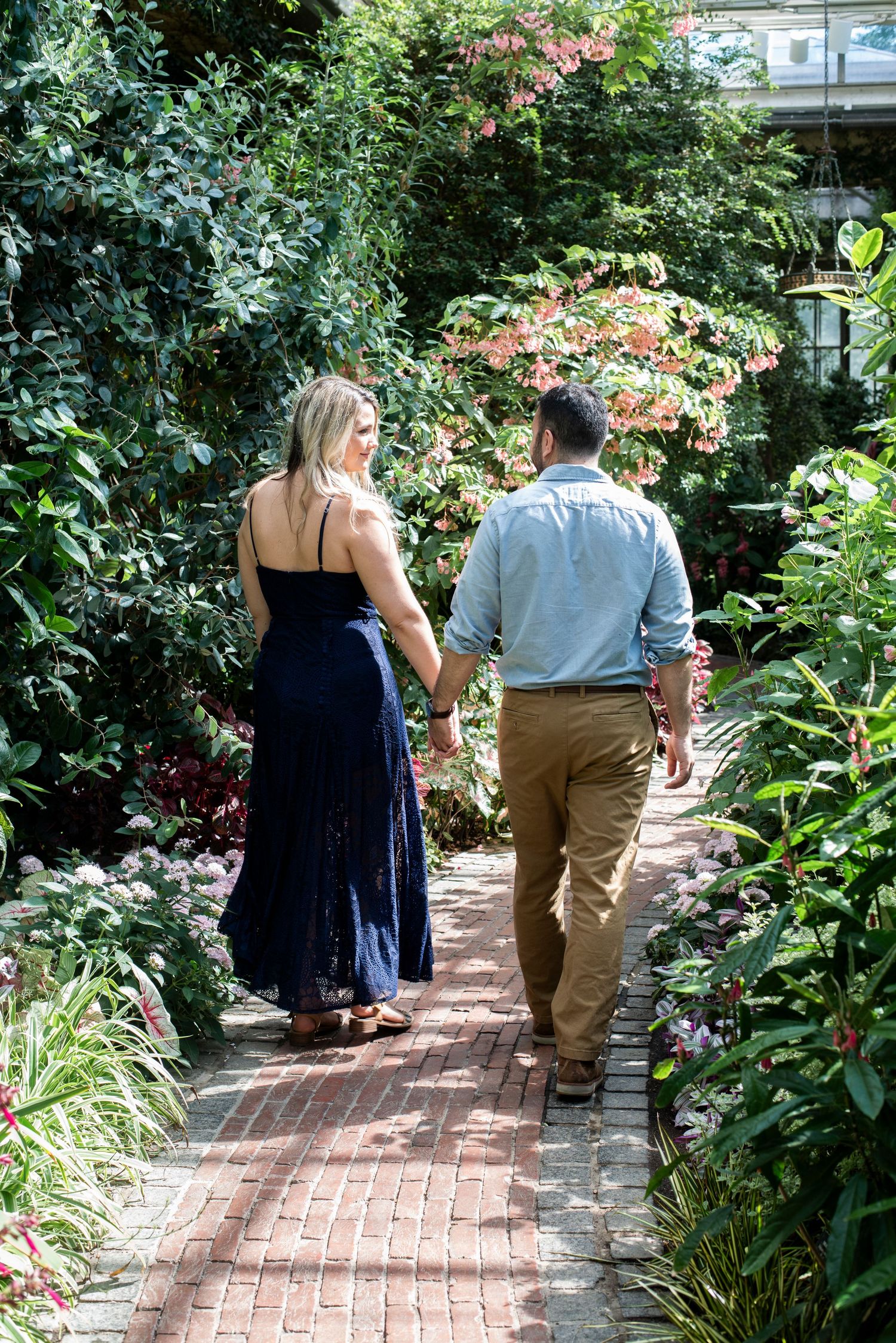 Engaged couple holds hands as they walk on a greenery-lined path during their engagement shoot at Longwood Gardens in Pennsylvania.