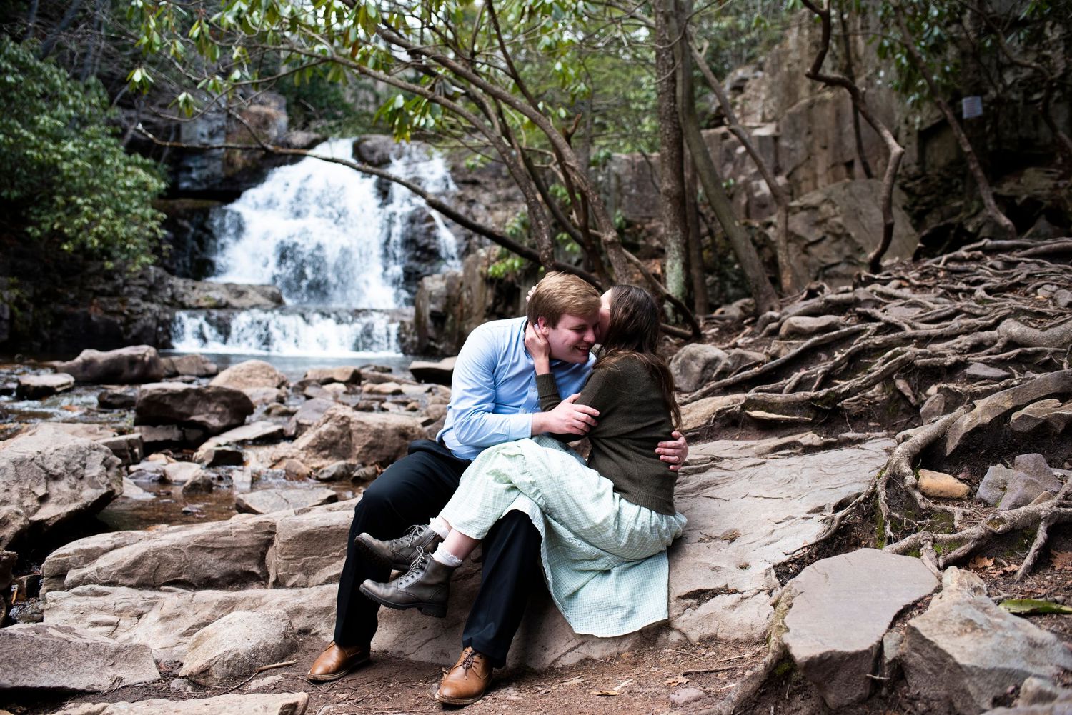 Couple sits on the rocks kissing in front of the waterfall during engagement shoot at Hawk Falls in Pennsylvania.
