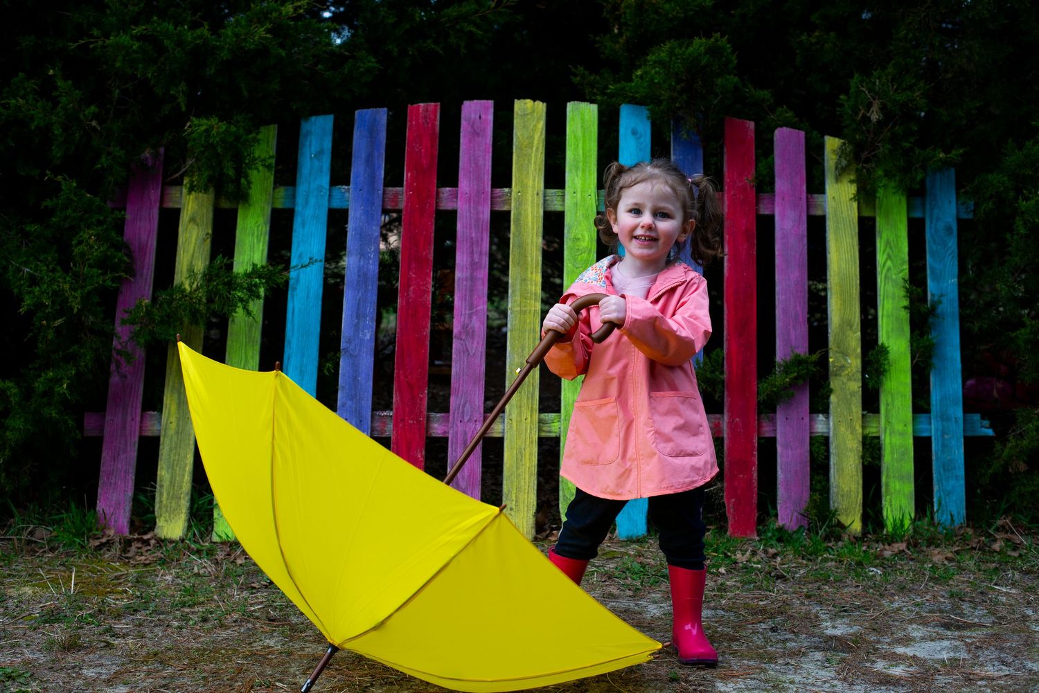 Young girl in a pink raincoat holds a yellow umbrella in front of a rainbow painted fence.