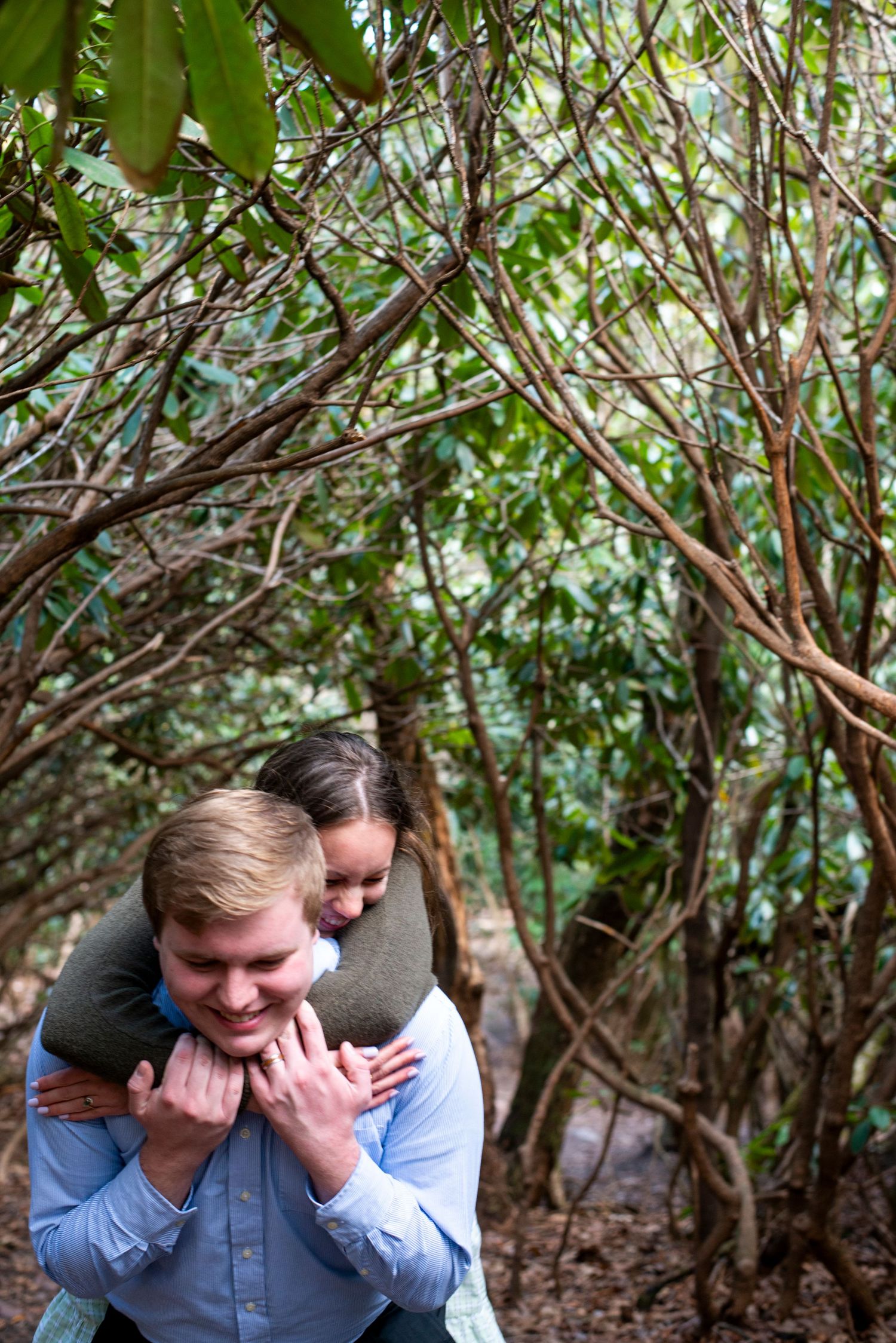 Bride jumps on groom's back under a thicket of trees during engagement shoot at Hawk Falls in Pennsylvania.