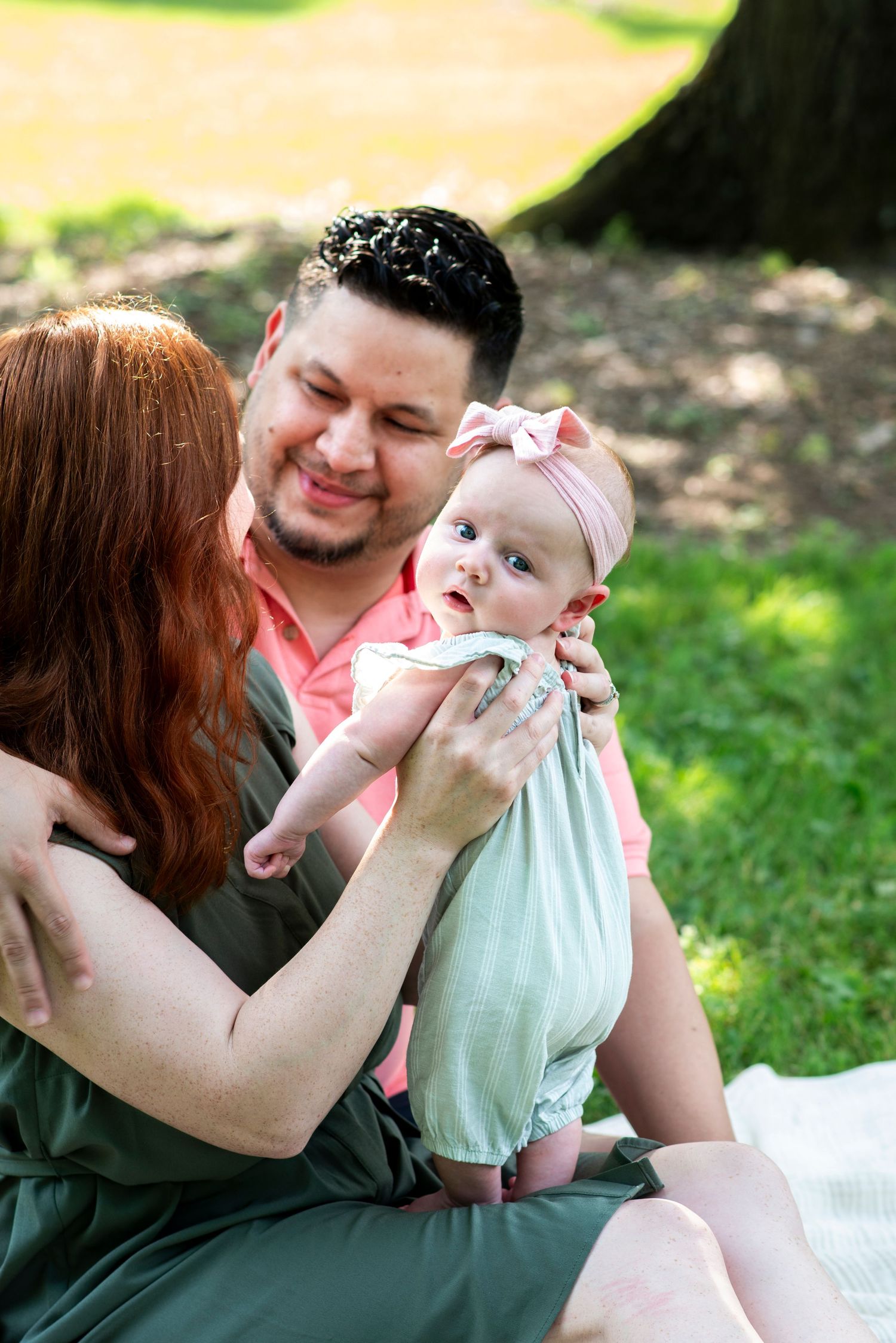 Family of three portrait in the grass, mom and dad smile at each other while baby stands on their lap.