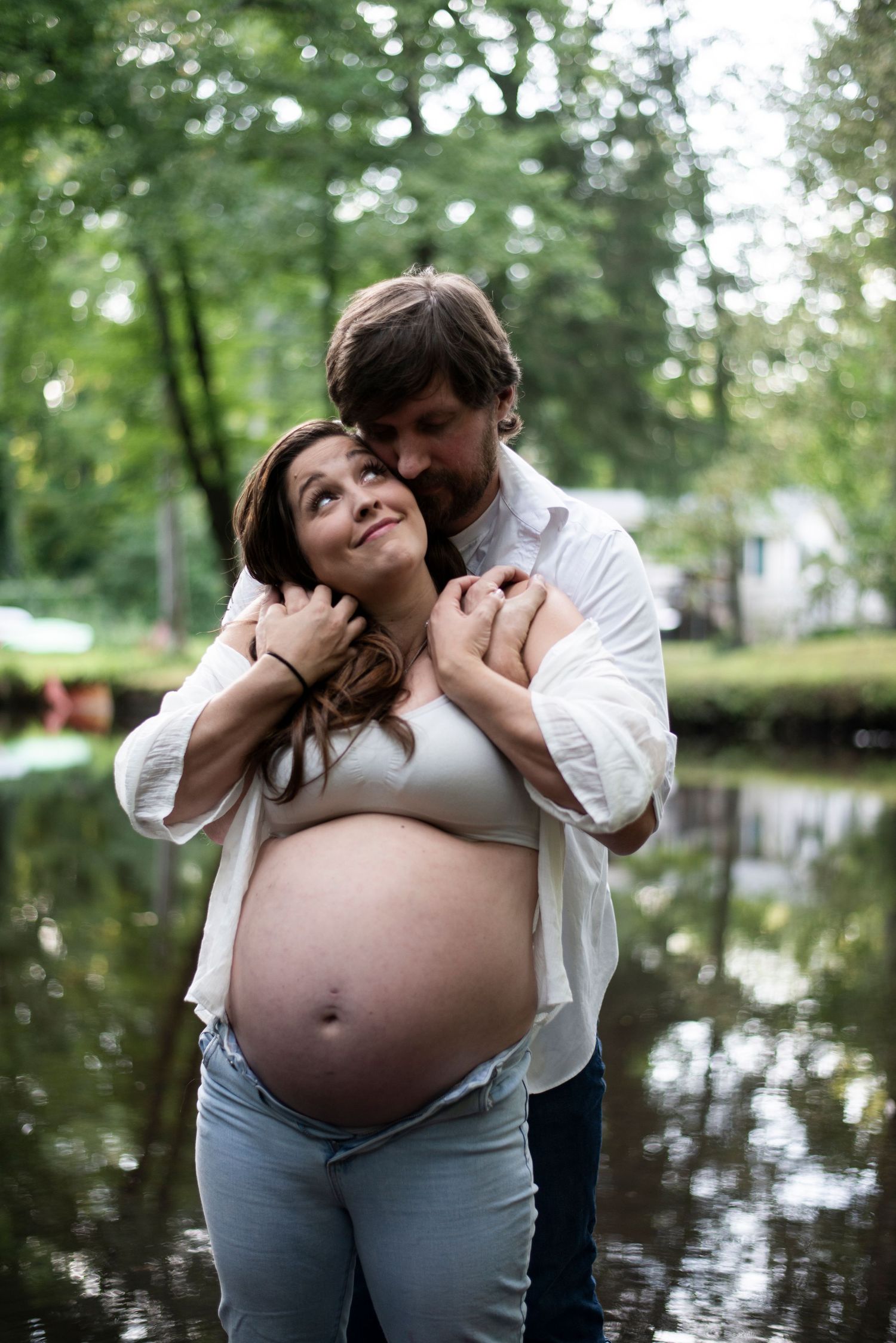 Man kisses his pregnant wife's cheek from behind as they stand in a creek in New Jersey. She wears jeans and a white shirt showing her belly