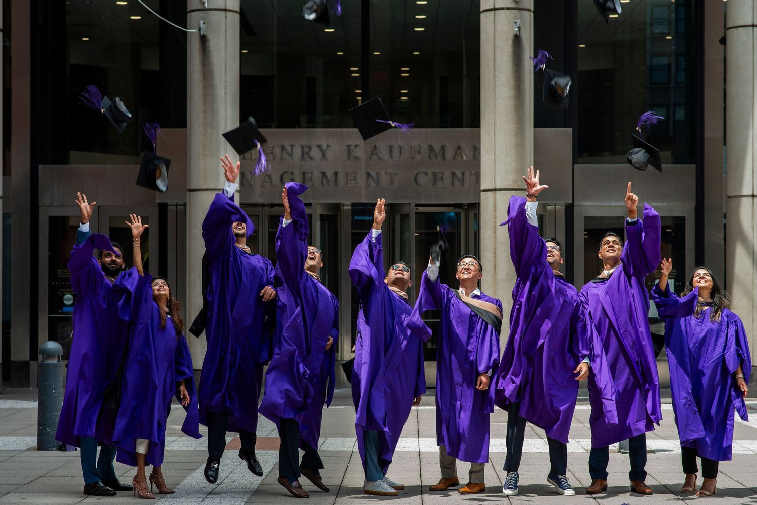 Group of nine NYU Stern business school graduates in purple robes toss their graduation caps into the air in New York City.