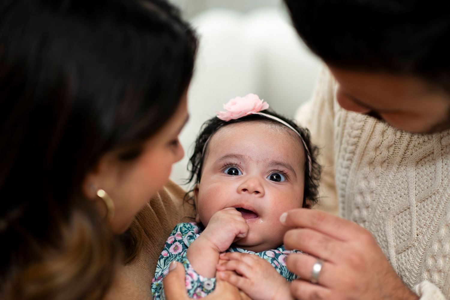 Baby girl looks up at the camera from between her parents as they look down at her and dad gently pinches her cheek.