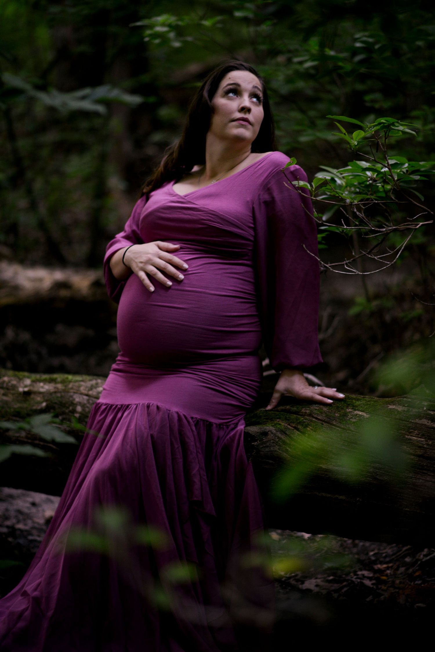 Beautiful pregnant woman in a dark mauve dress reclines in the forest, looking back over her shoulder.
