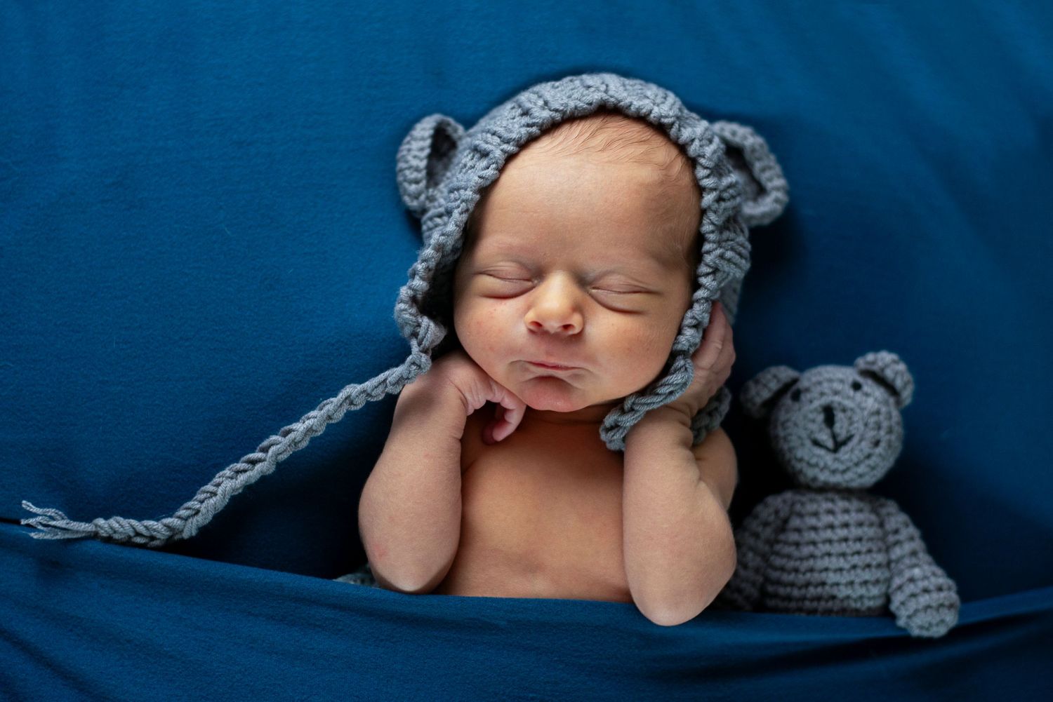Portrait of newborn baby with gray bear hat and matching bear stuffed animal on navy background.